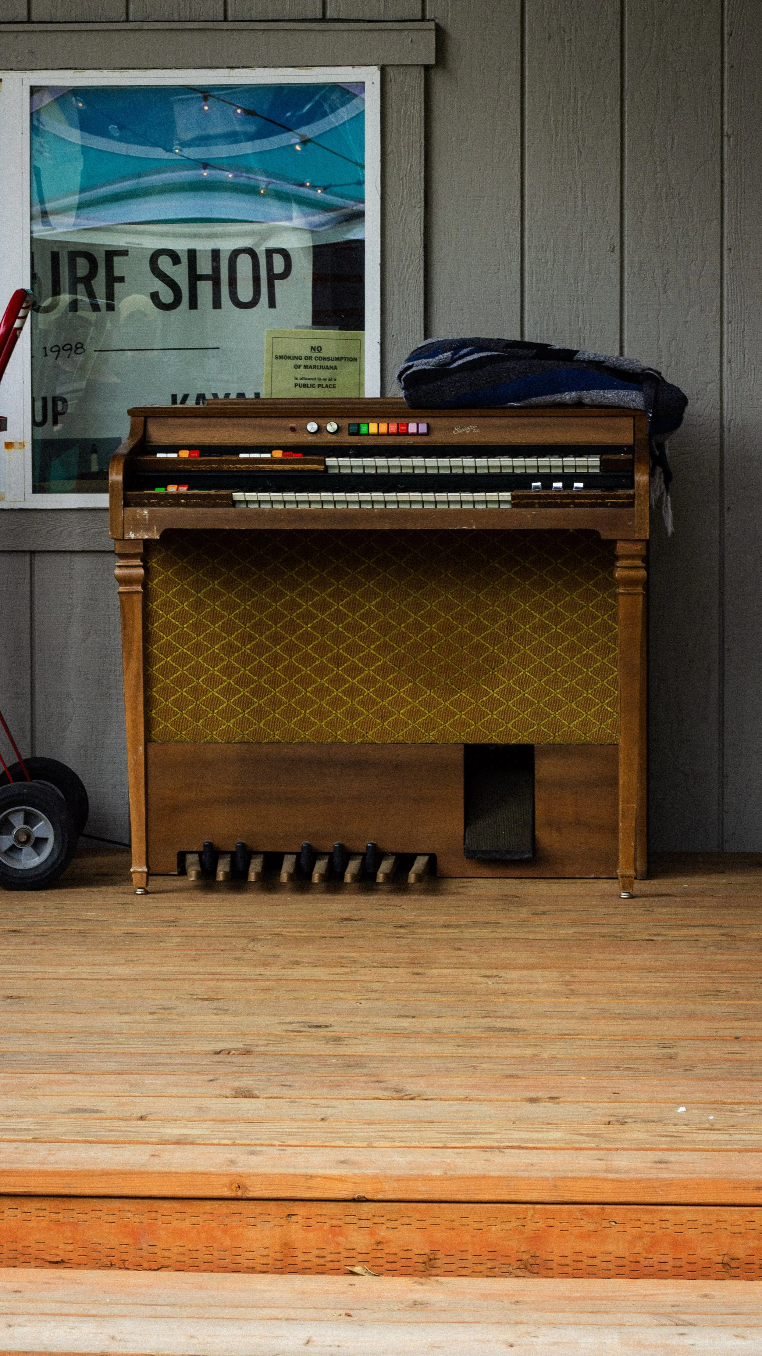 Vintage wooden organ with yellow patterned speaker cover, placed on wooden floor in a room with gray panel walls. A striped blanket or cloth rests on top of the organ. To the left, a small red-handled tool or object is visible. A window behind the or