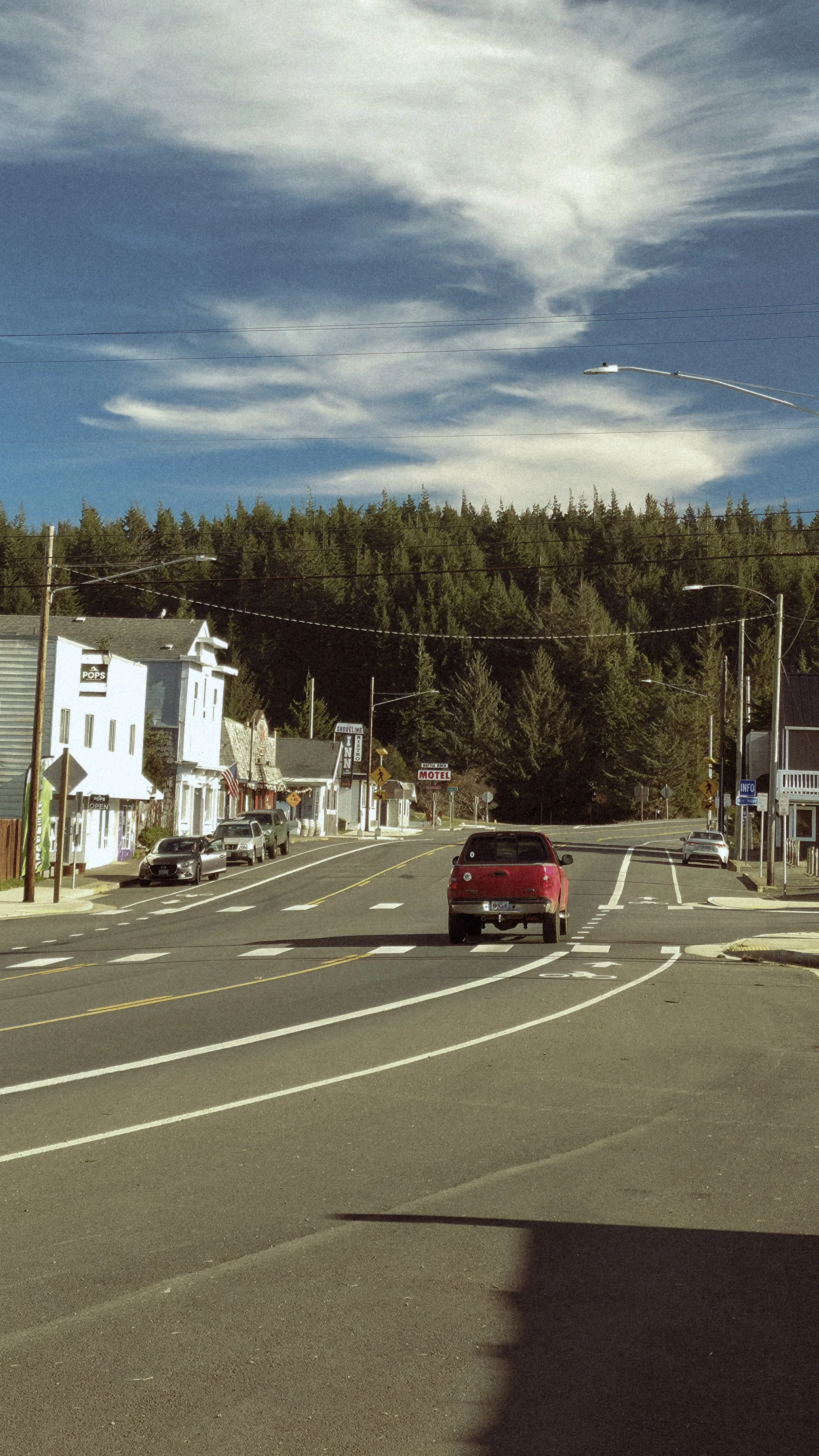 A small town street scene with parked cars, storefronts, and a forested hillside in the background under a partly cloudy sky.