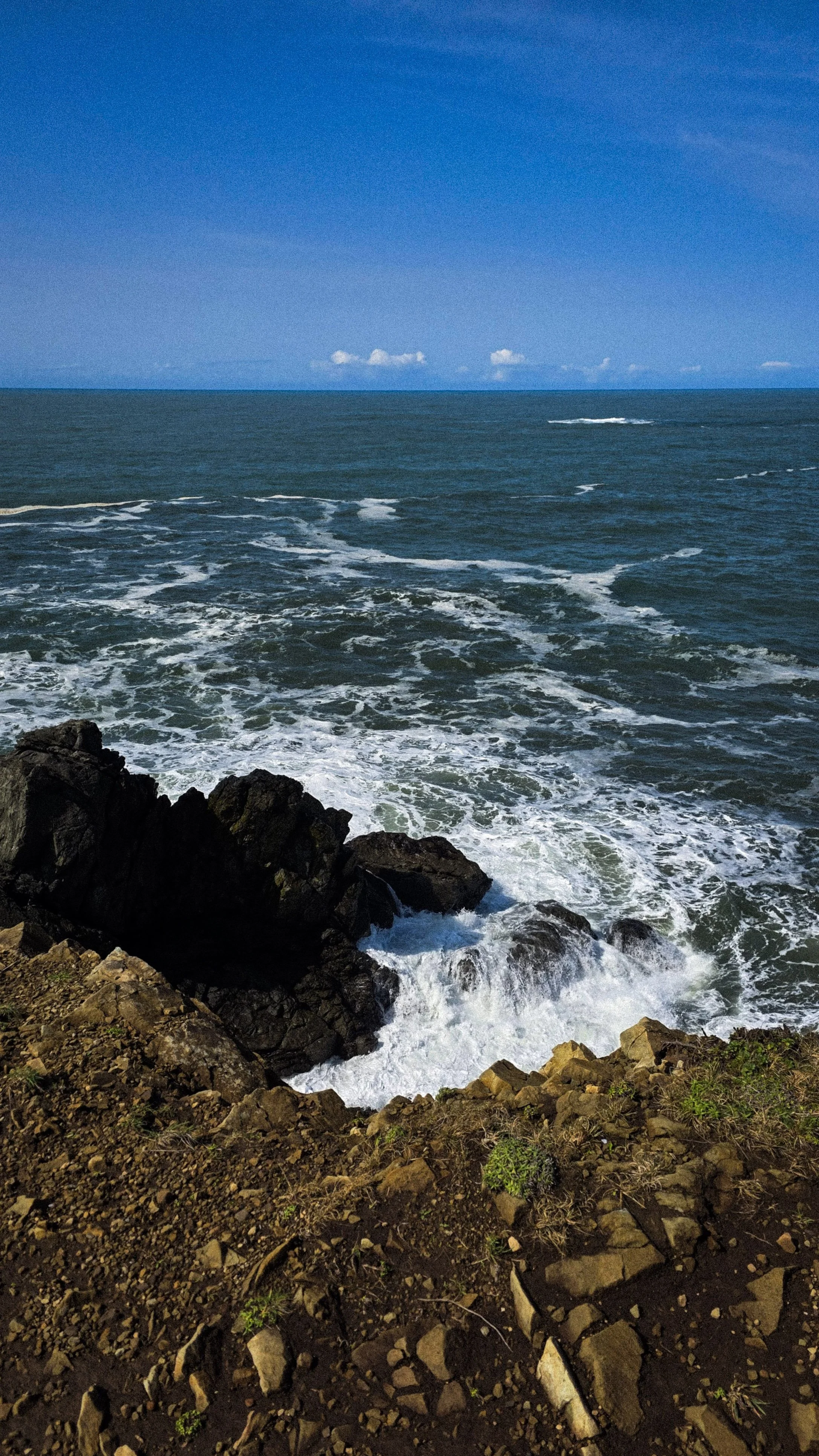 A rocky shoreline with the ocean and blue sky in the background, and some small clouds on the horizon.