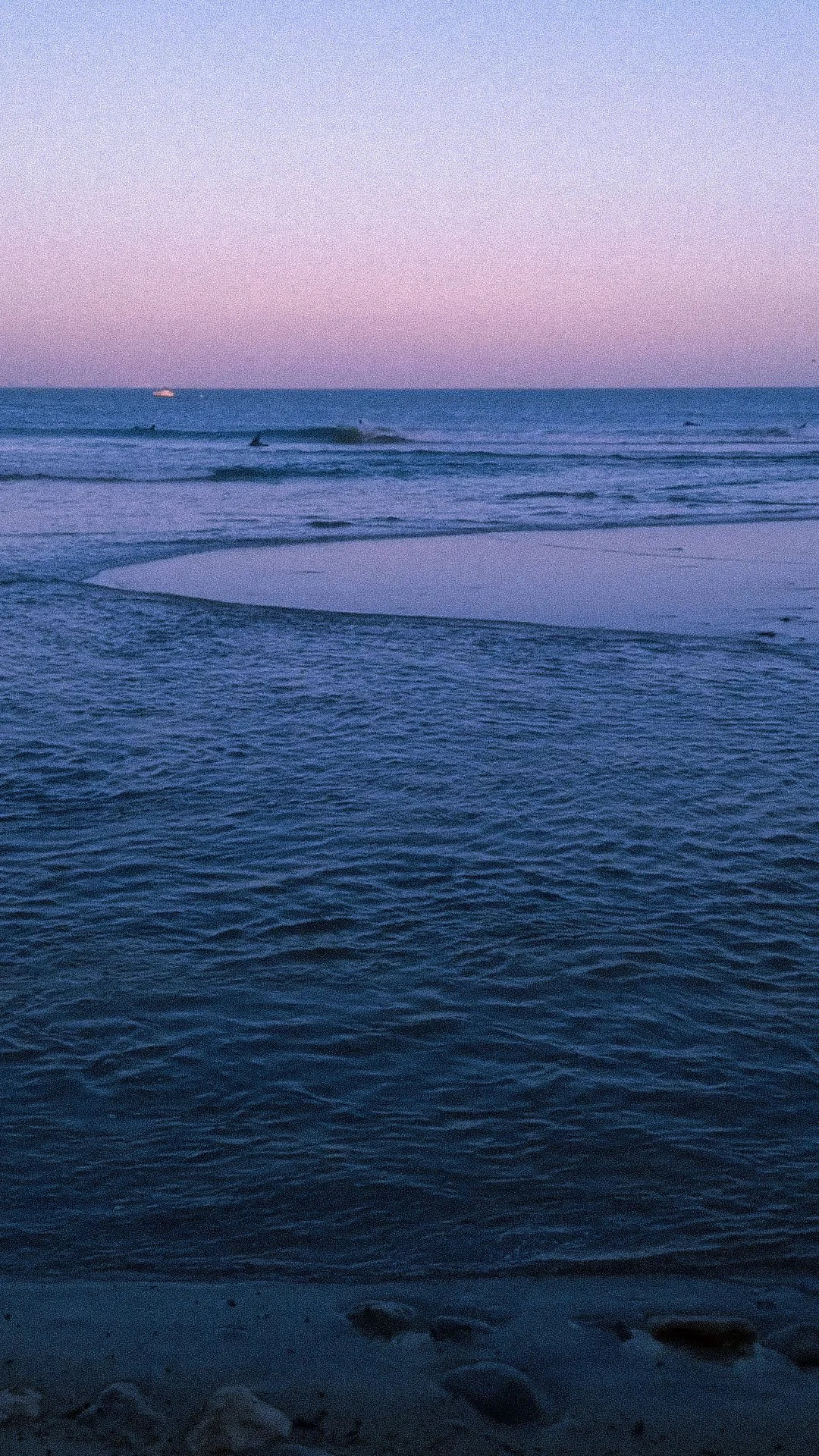 View of the ocean at sunset with gentle waves, a boat in the distance, and rocks on the sandy beach in the foreground.