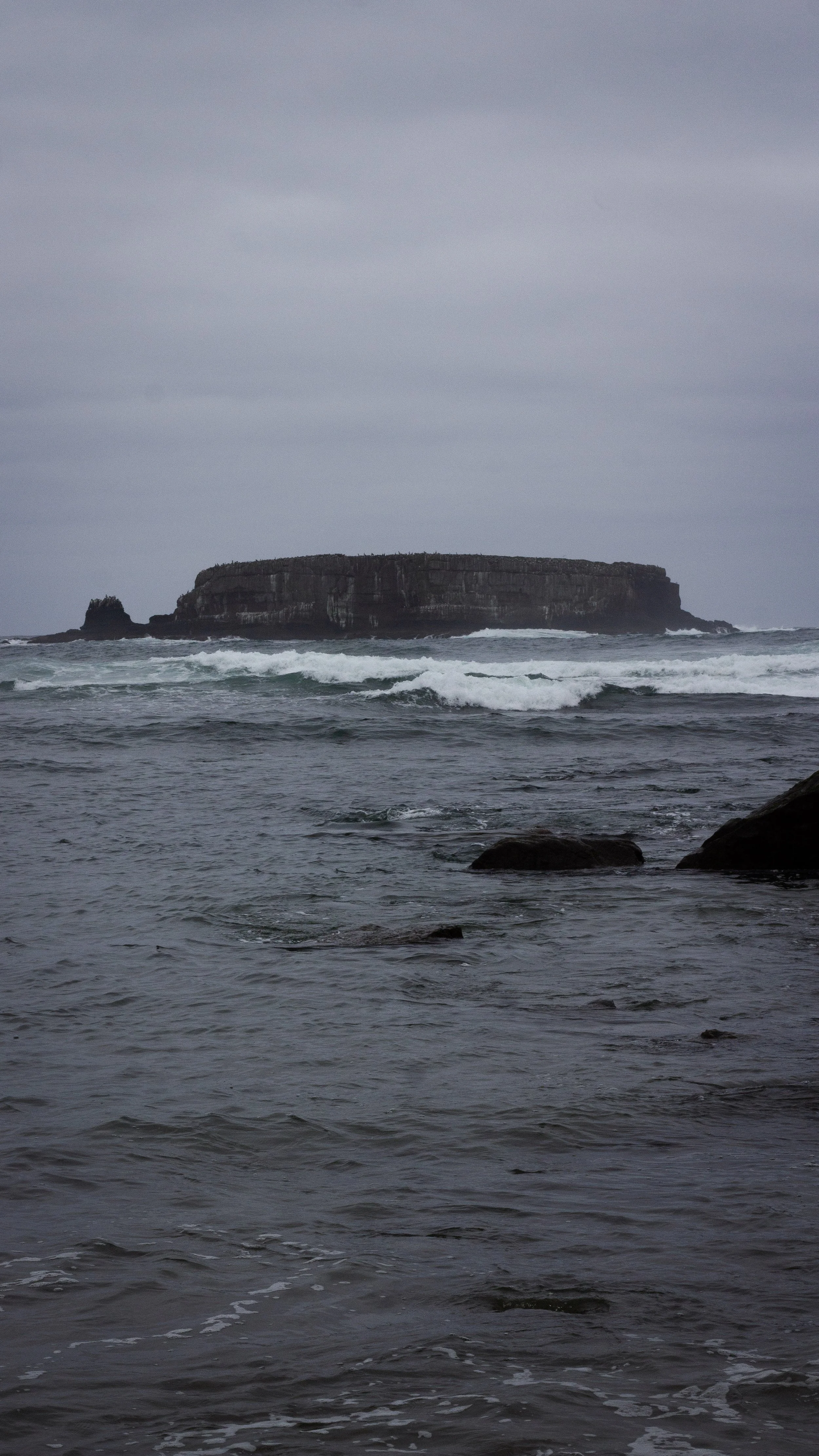 Ocean waves crashing against rocks with a large flat-topped island or plateau in the distance under an overcast sky.