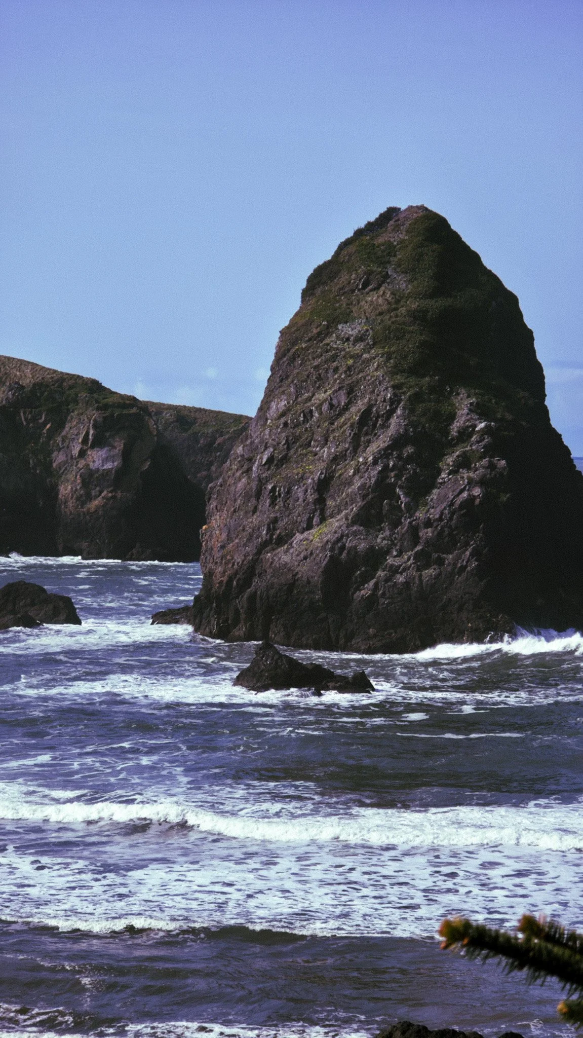 Large rocky sea stack near the coastline with smaller rocks in the water and a blue sky overhead.