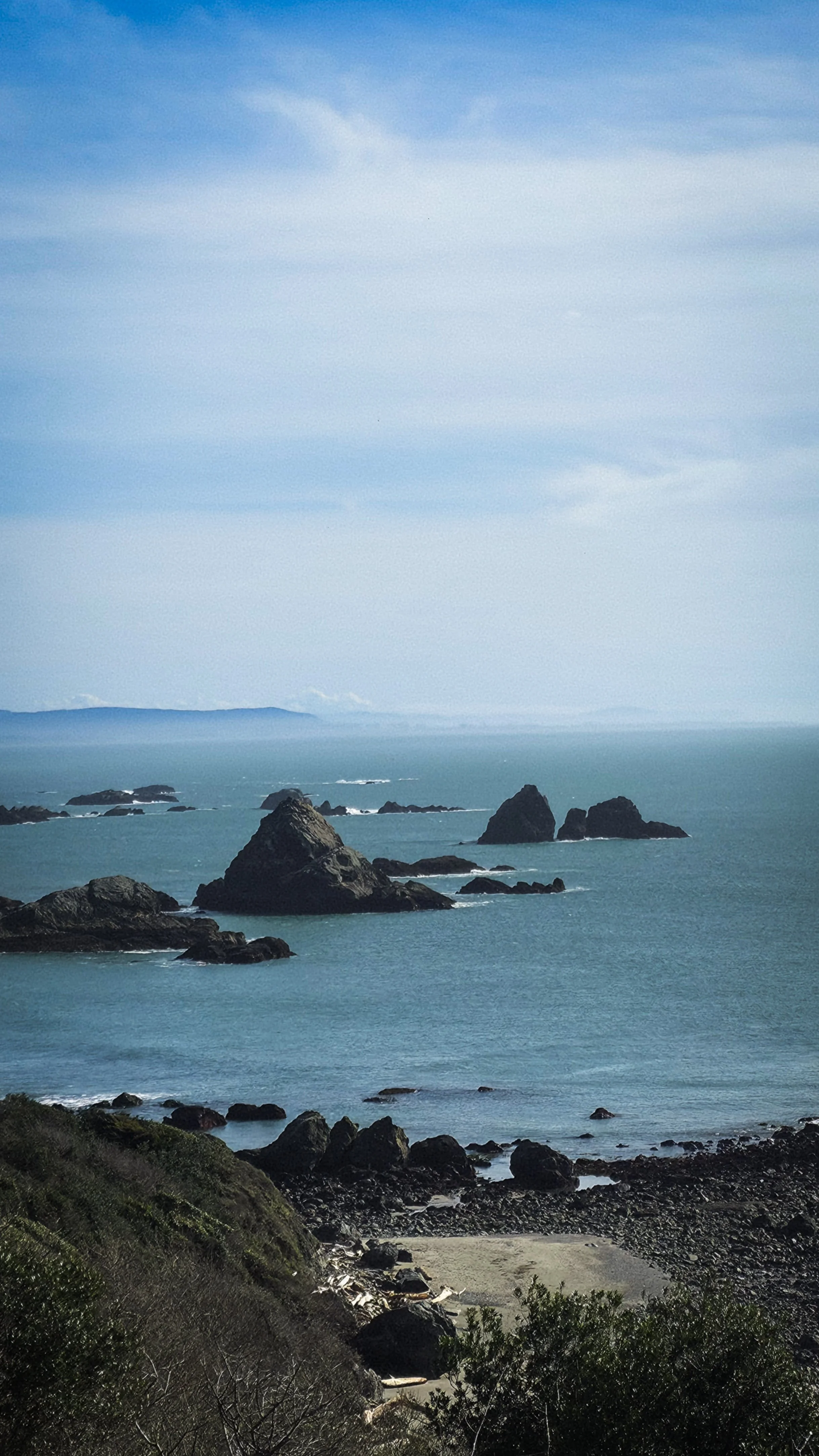 Ocean coastline with rocky outcroppings and a sandy beach in the foreground. The sky is partly cloudy with a mix of blue and white clouds. Distant mountains or hills can be seen across the water.