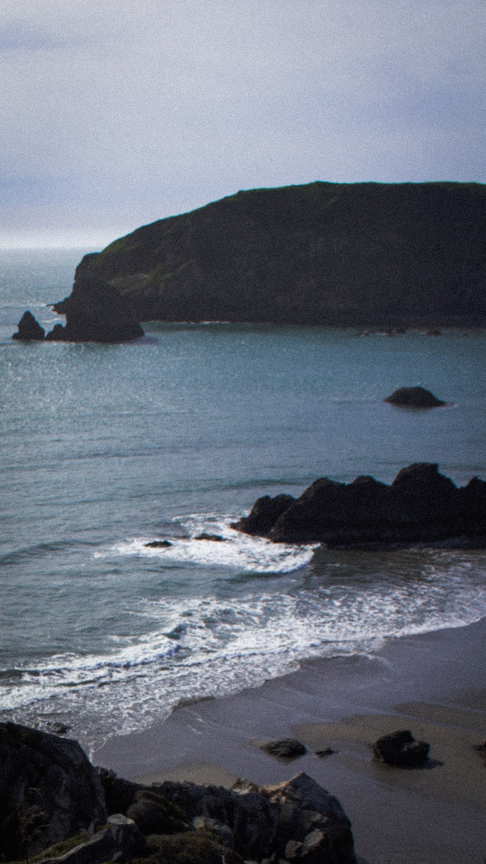 A rocky coastal landscape at dusk with a large hill or small mountain in the background, calm ocean waters, and waves gently hitting the shoreline.
