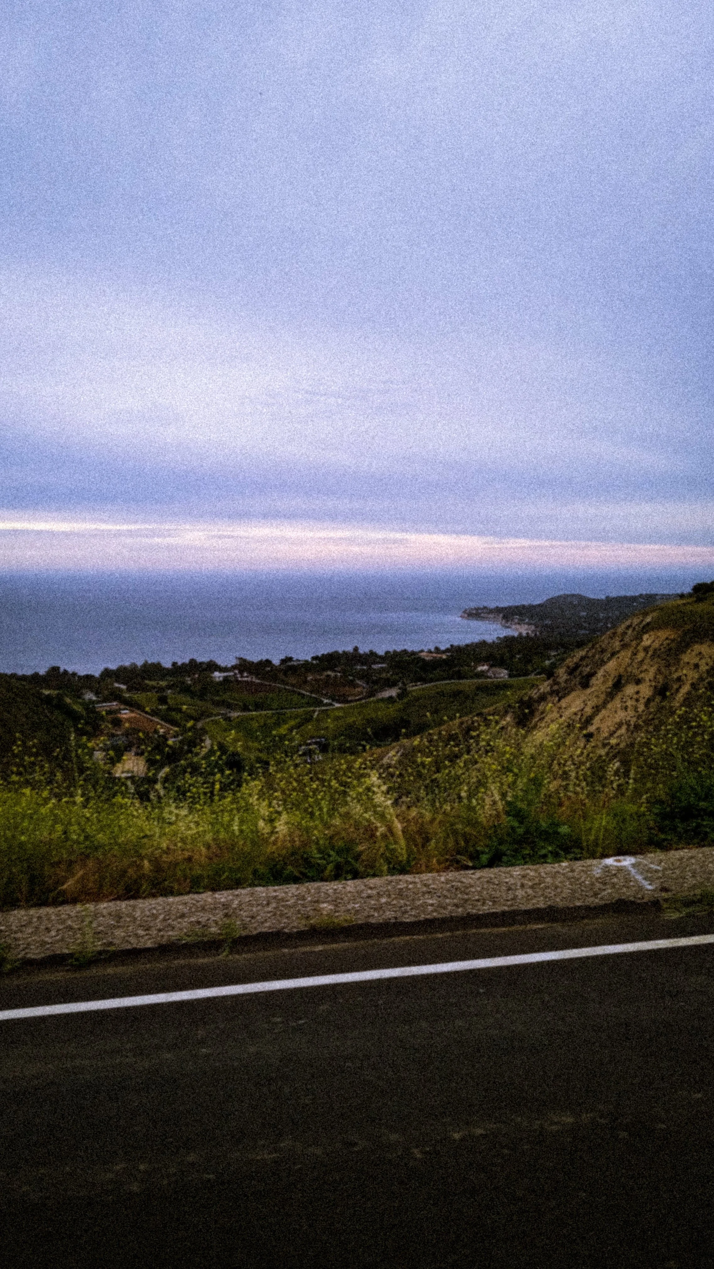 Scenic view of coastline and ocean during dusk, with cloudy sky and hilly landscape in foreground, taken from the side of a road.