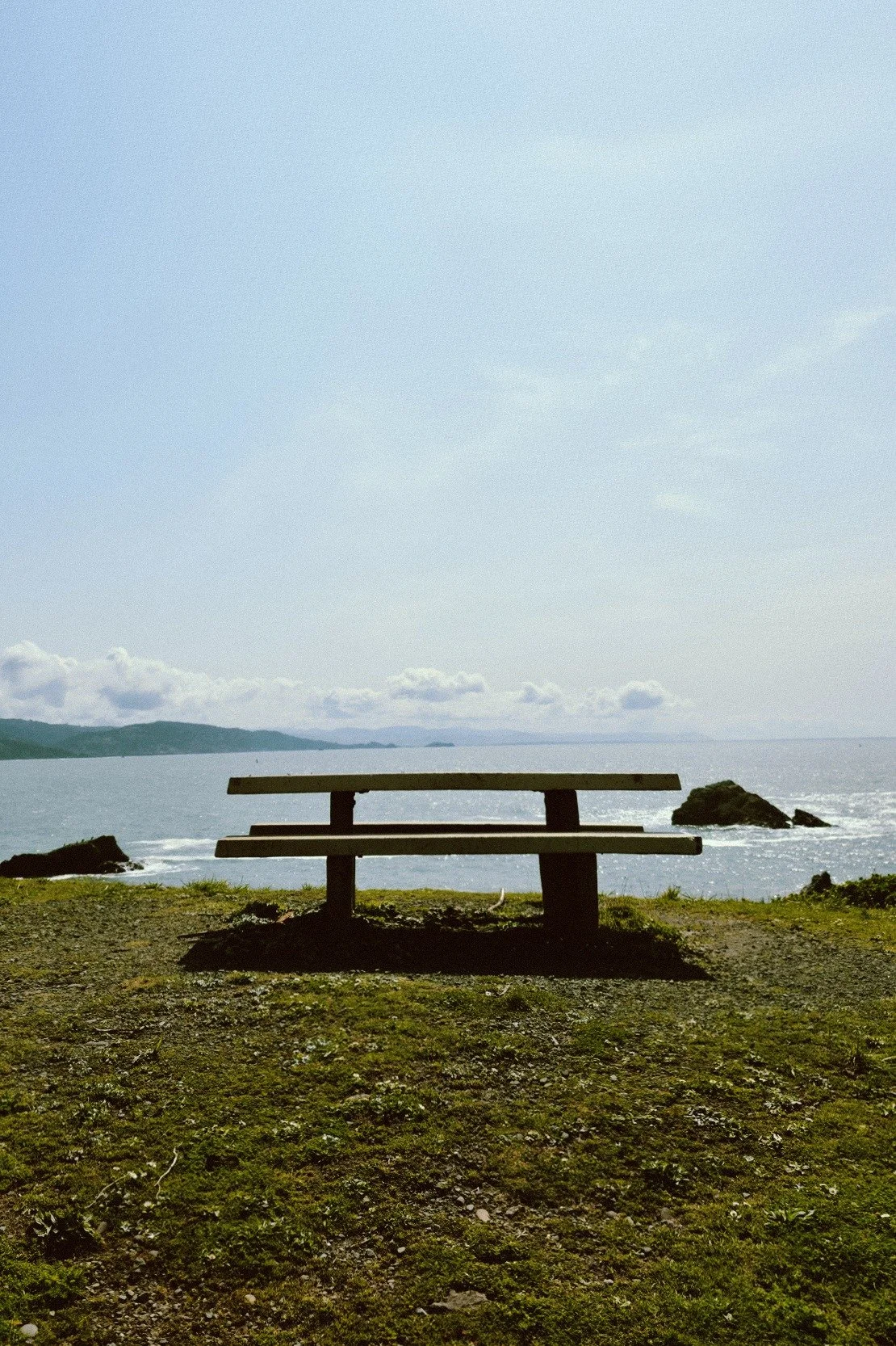 A wooden bench on a grassy area facing the ocean with rocks and hills in the background under a partly cloudy sky.