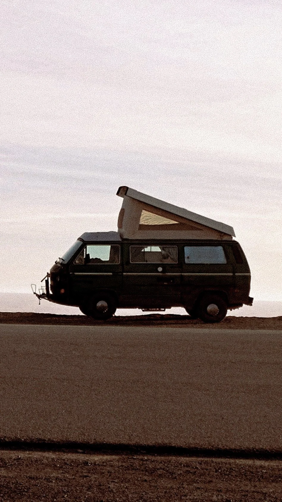A black camper van with a pop-up roof parked on a flat terrain during sunset or sunrise, with a pastel sky in the background.