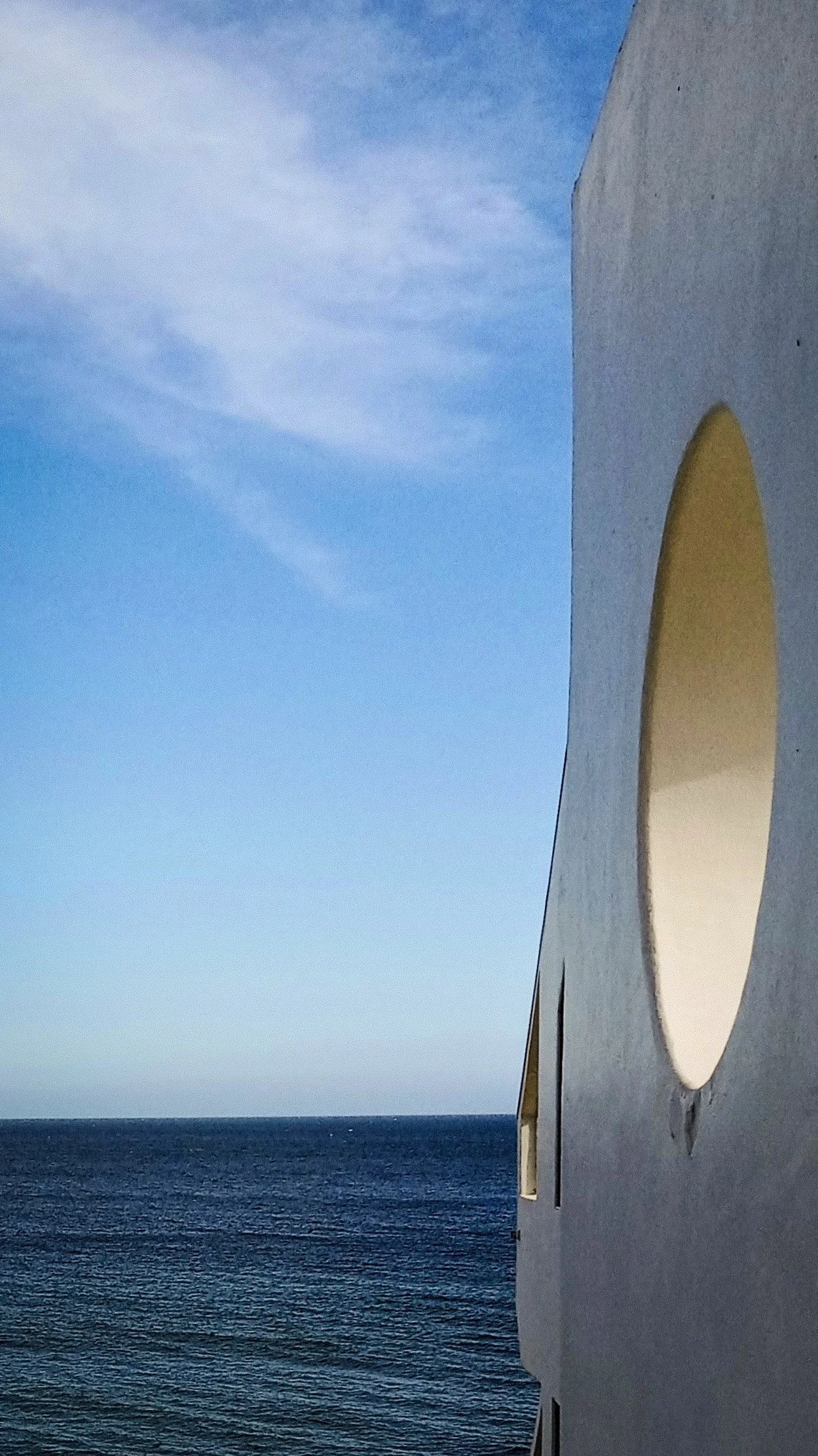 Part of a white building with a large round window on the right side, overlooking the ocean under a partly cloudy sky.