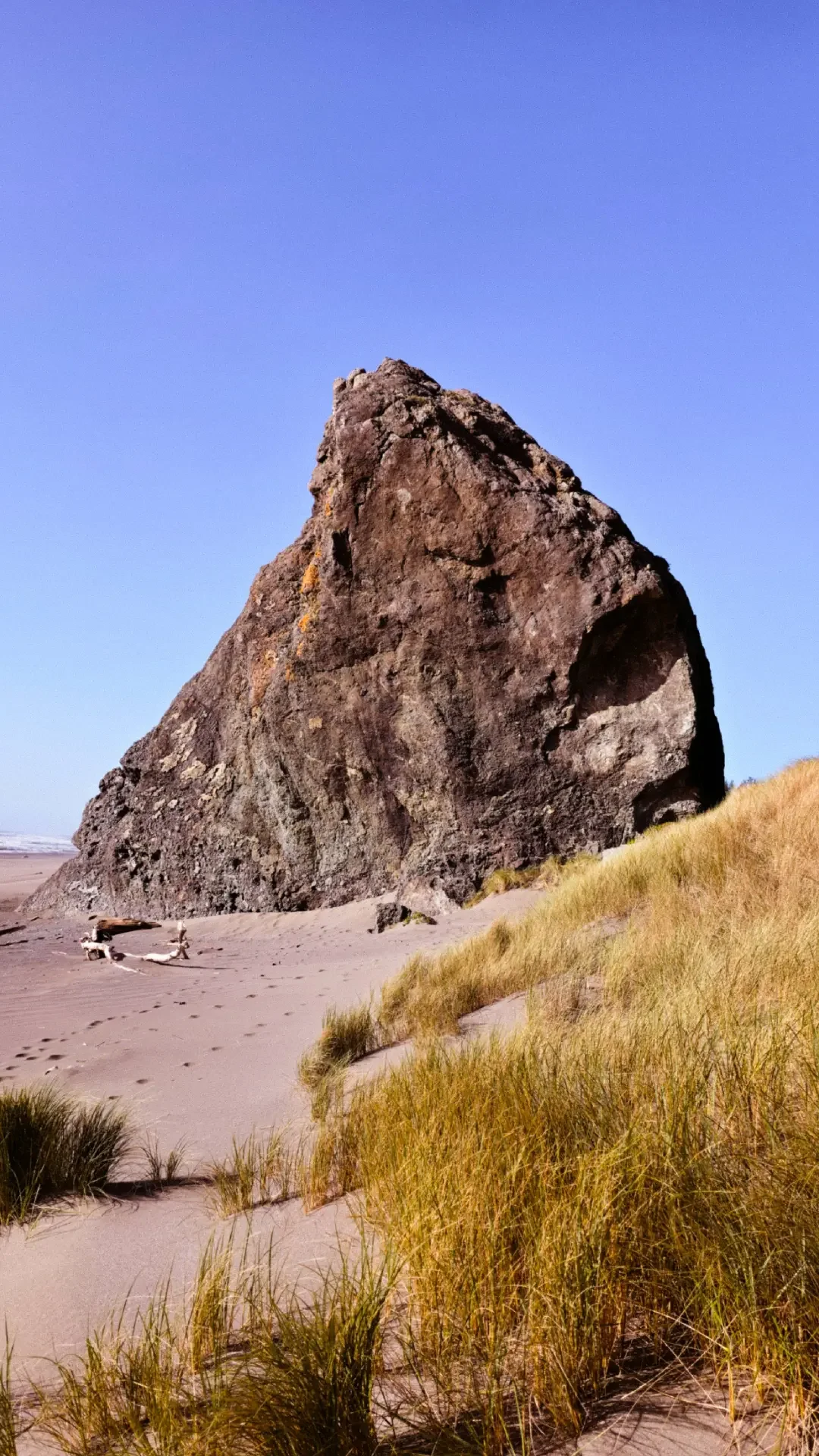A large, triangular rock formation on a sandy beach with grassy dunes in the foreground and a clear blue sky.