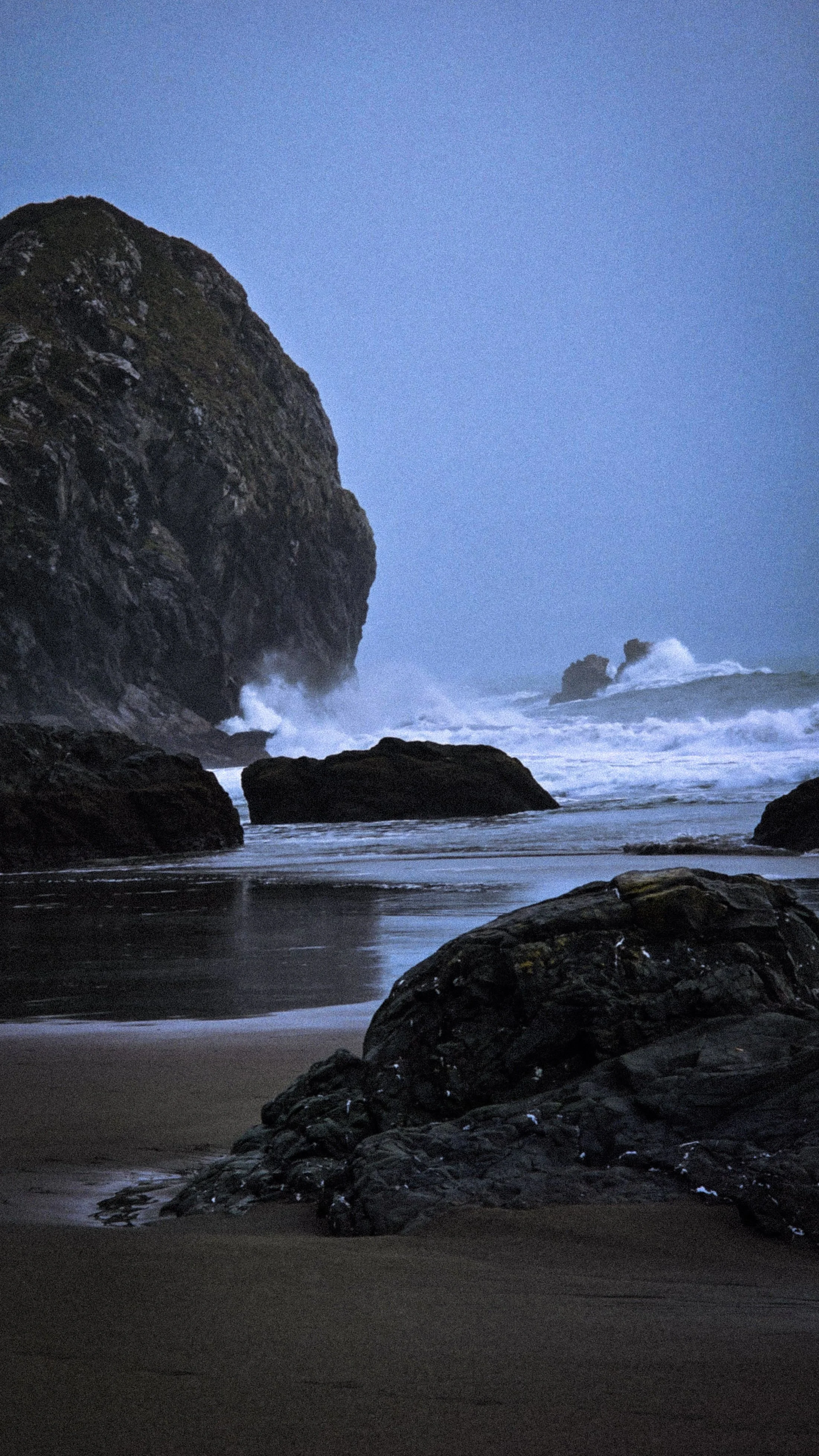 Dark rocky shoreline with large rocks near the water, waves hitting rocks, and a large cliff on the left side during twilight or dusk.