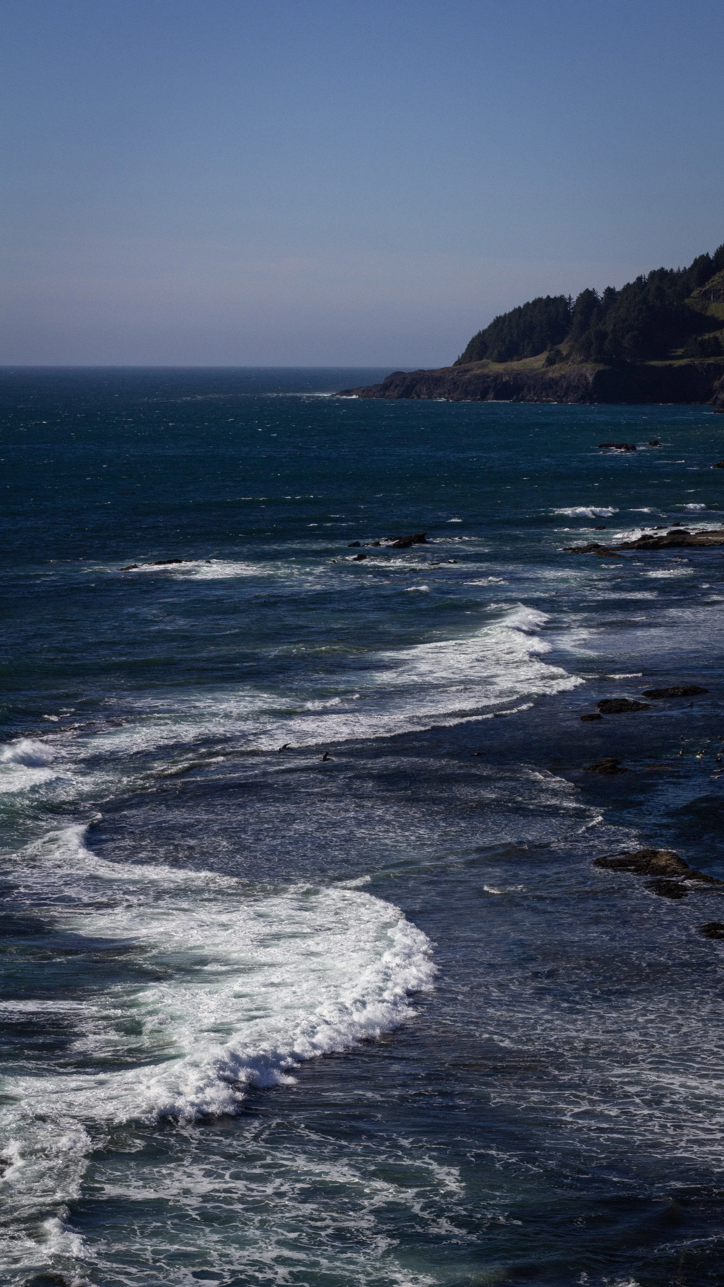 Ocean waves crashing against a rocky shoreline with green hills in the background under a clear blue sky.