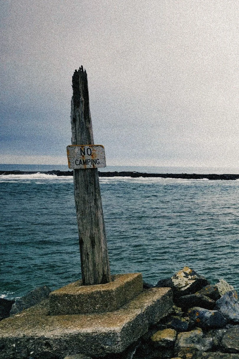 A weathered wooden pole with a 'No Camping' sign attached, situated on a concrete platform at the edge of the water, with rocks and the ocean in the background.
