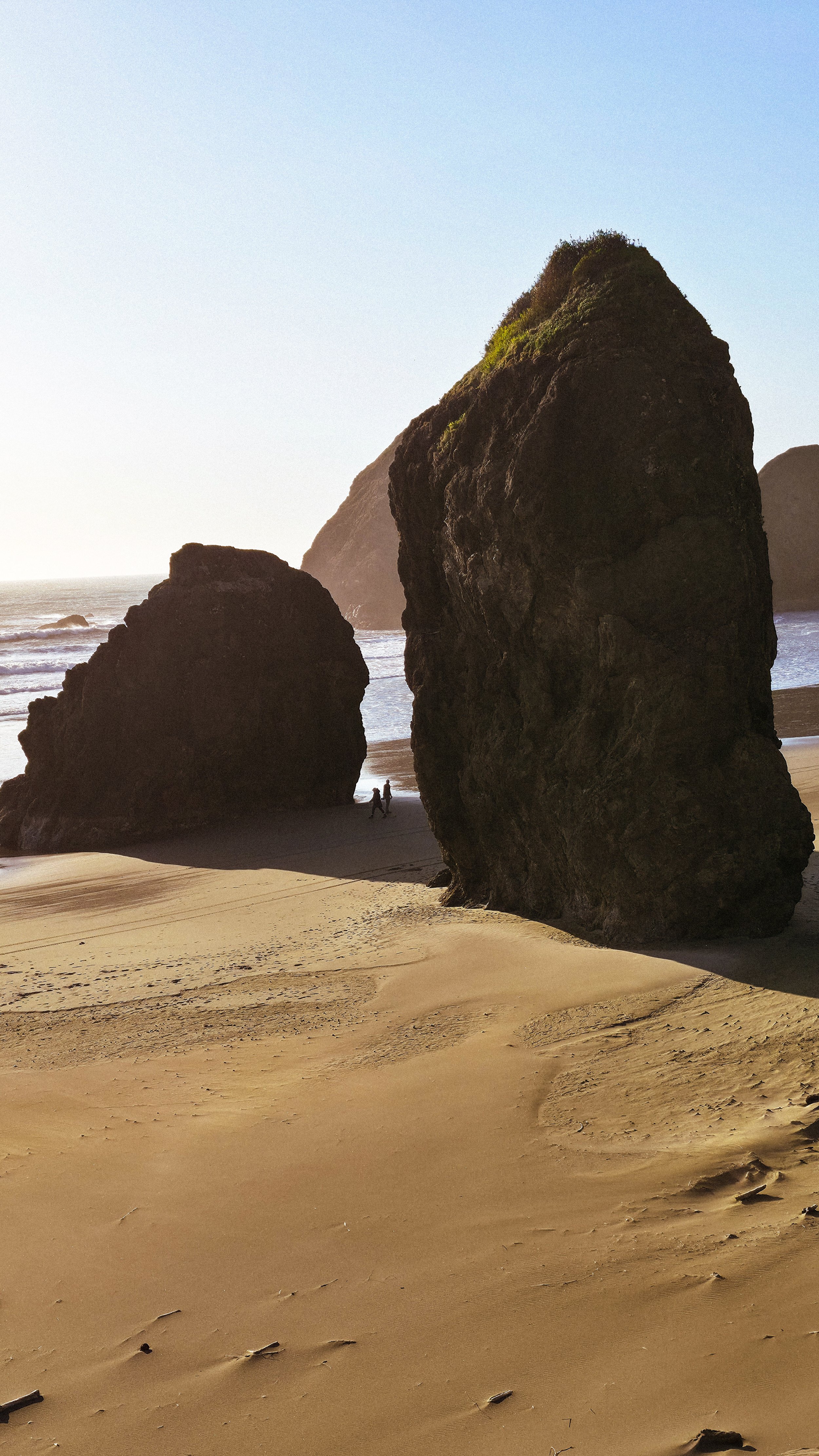 Large coastal rocks on sandy beach with two people walking near water and cliffs in the background, under clear sky.