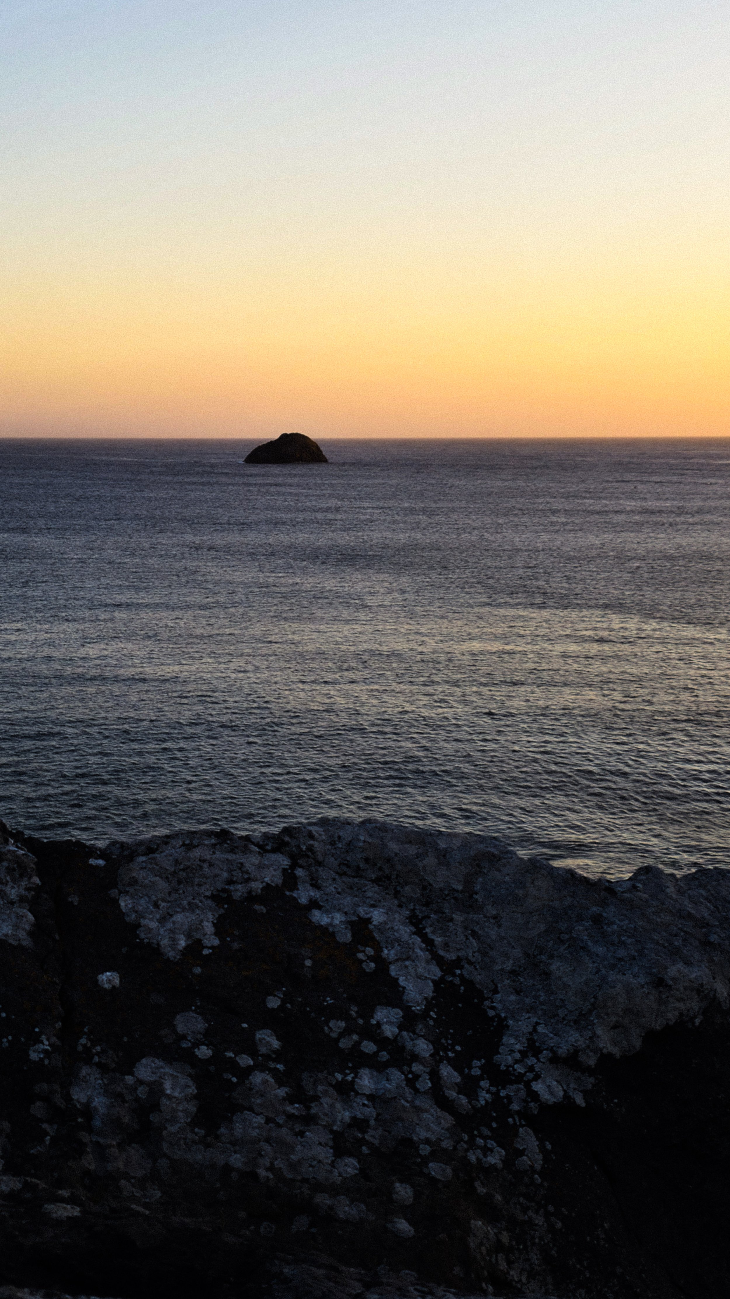 A sunset over the ocean with a large rock formation in the distance and dark rocks in the foreground.