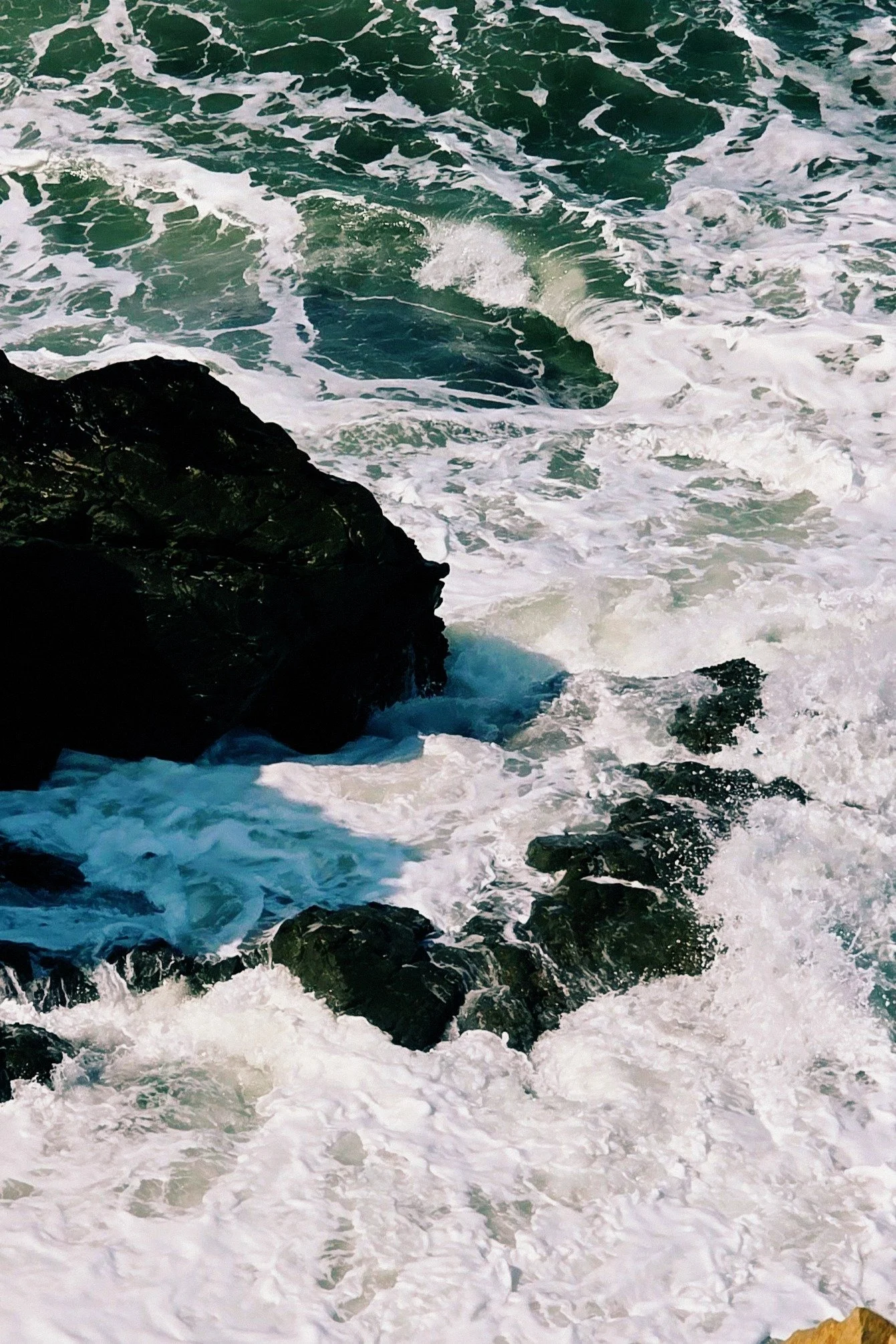 Ocean waves crashing against dark rocks along a shoreline, with white foam and churning water.