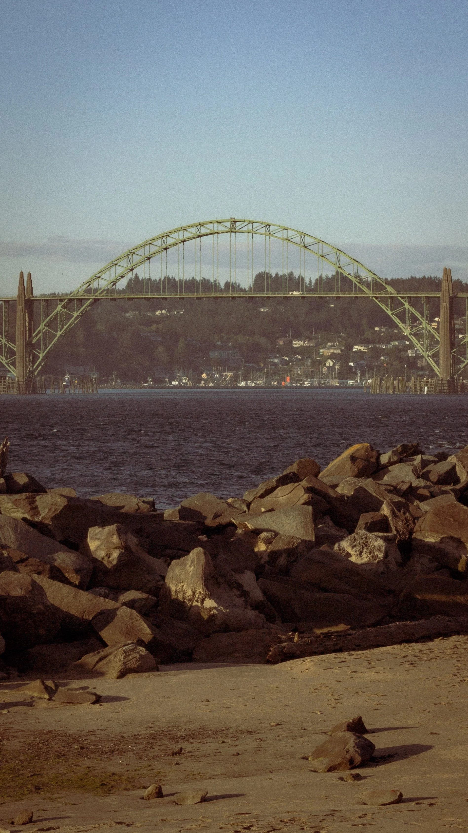 View of a large yellow arch bridge over water, with a forested hillside in the background and rocks along the shoreline in the foreground.