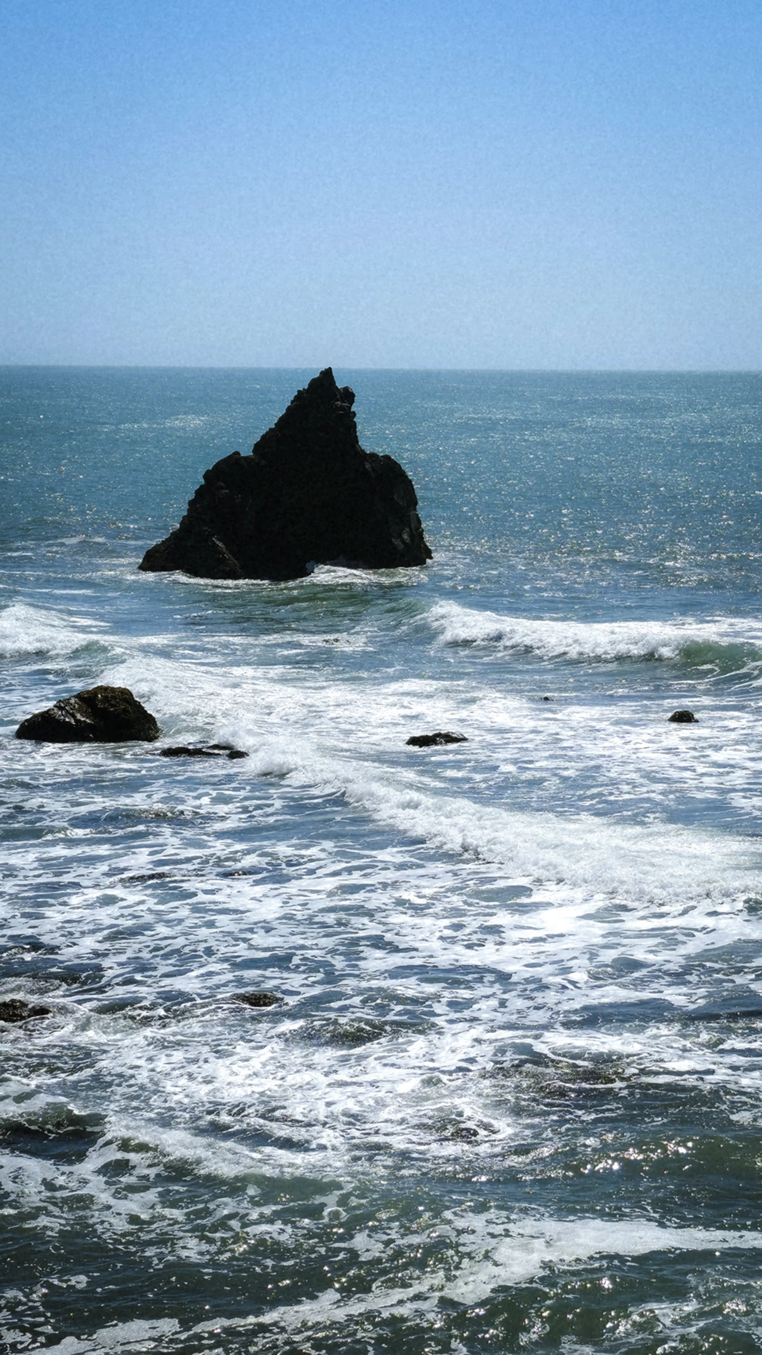 Large black rock formation in the ocean with waves crashing against it and smaller rocks nearby, under a clear blue sky.