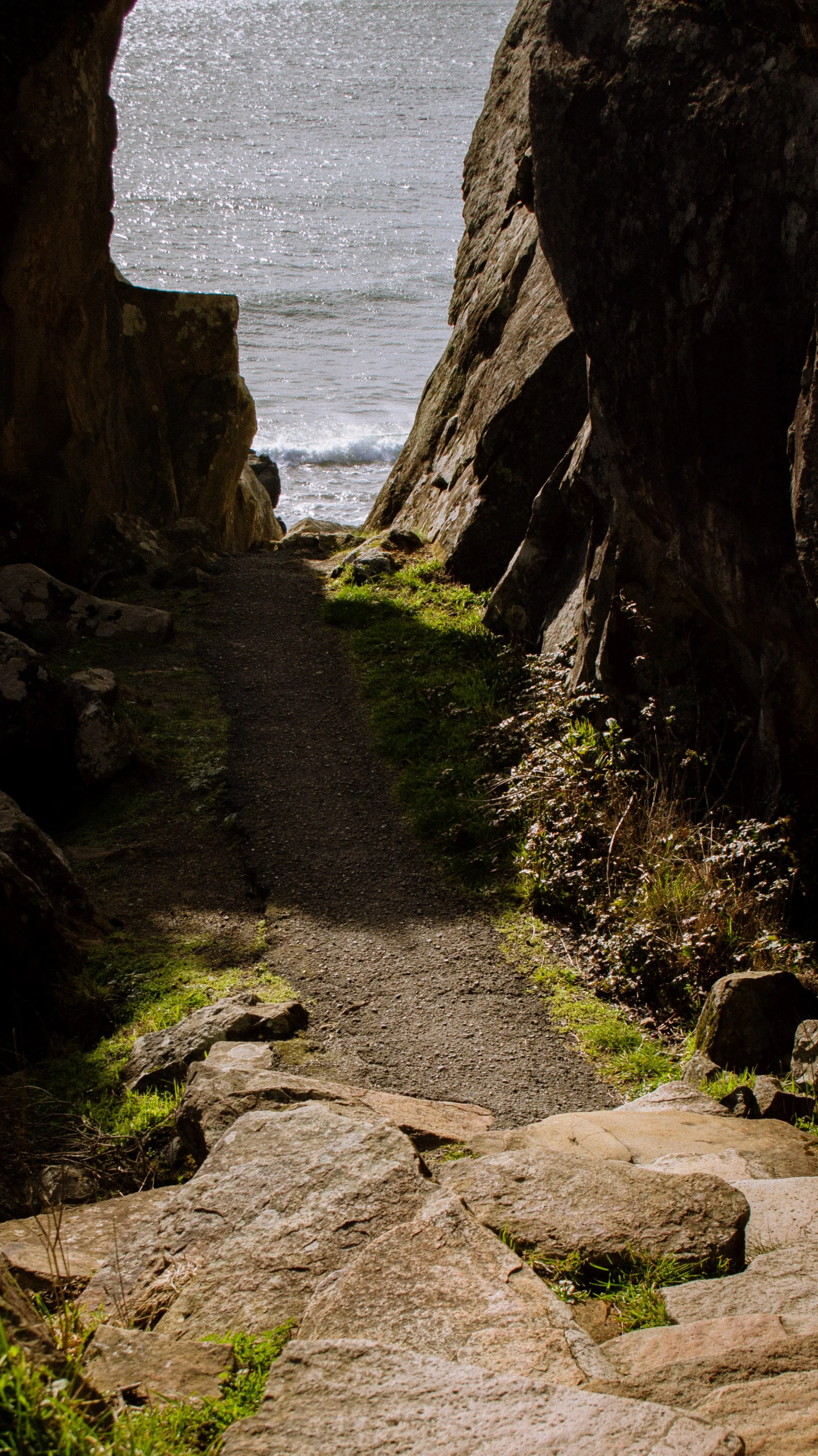 A narrow dirt trail between large rocks, leading towards the ocean with waves and sunshine in the background.