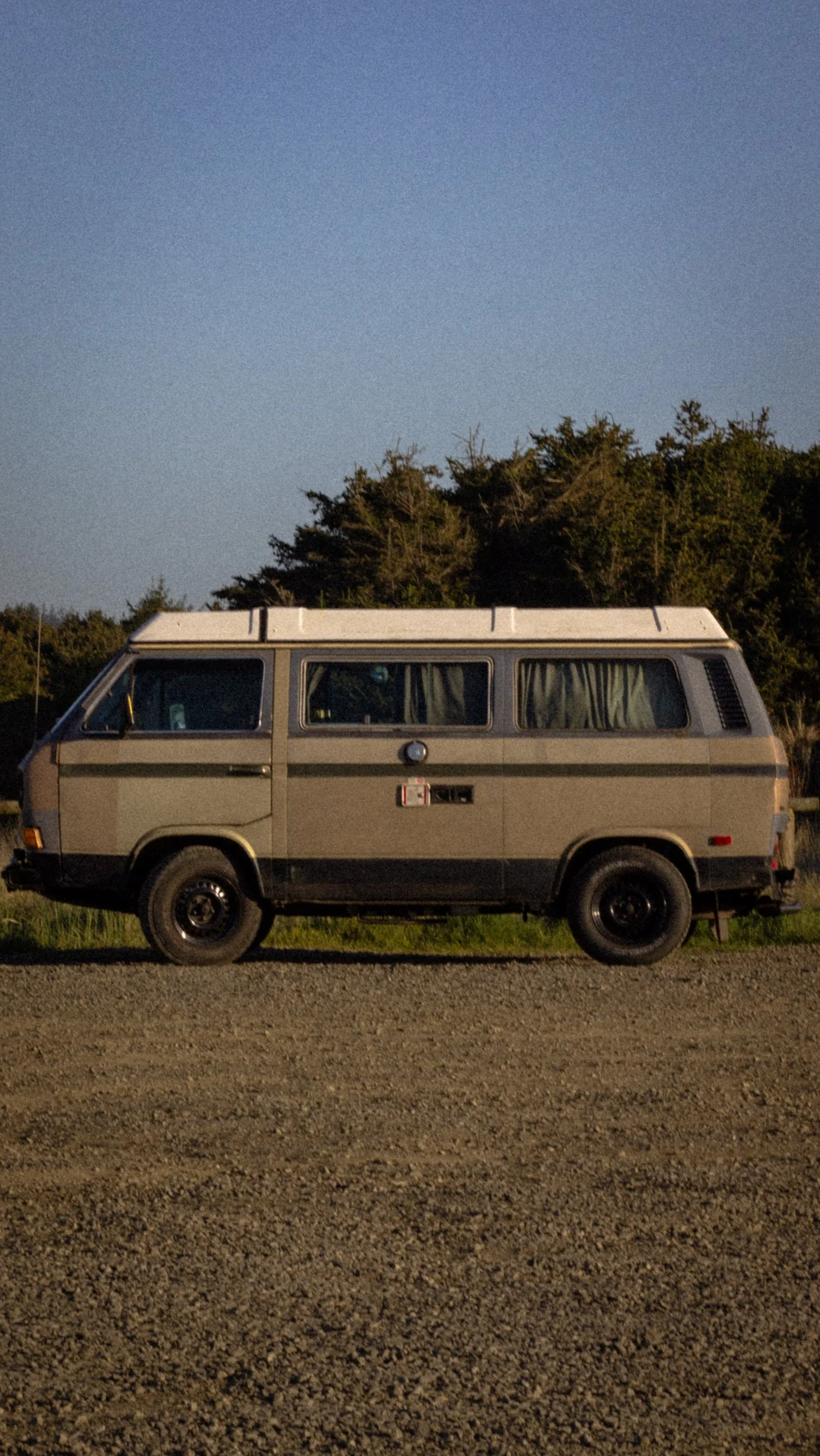A vintage beige van parked on a gravel road with trees and a blue sky in the background.