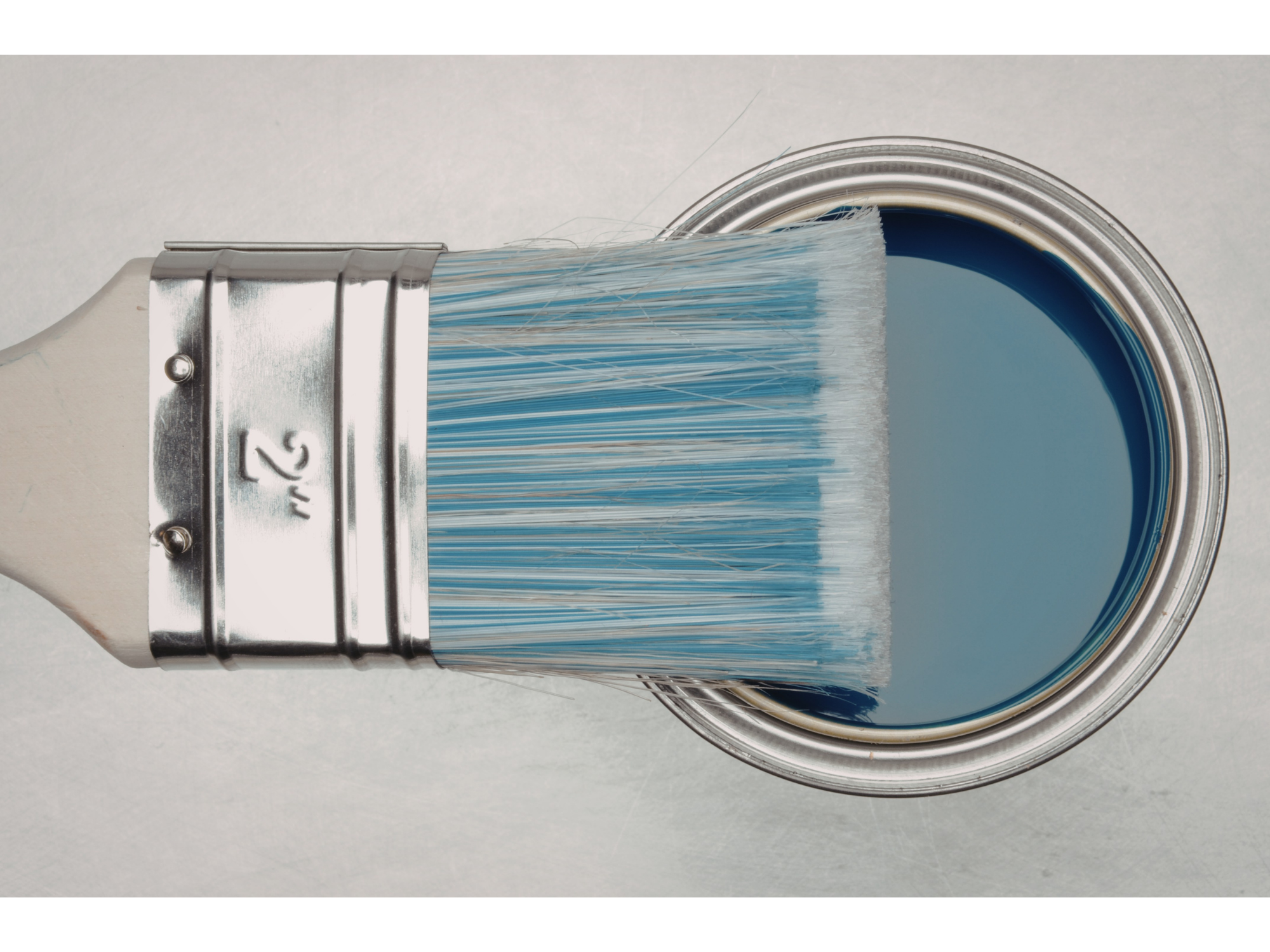 Close-up of an open paint can with blue paint and a paintbrush resting on the rim. The paintbrush has a metallic handle and white bristles.