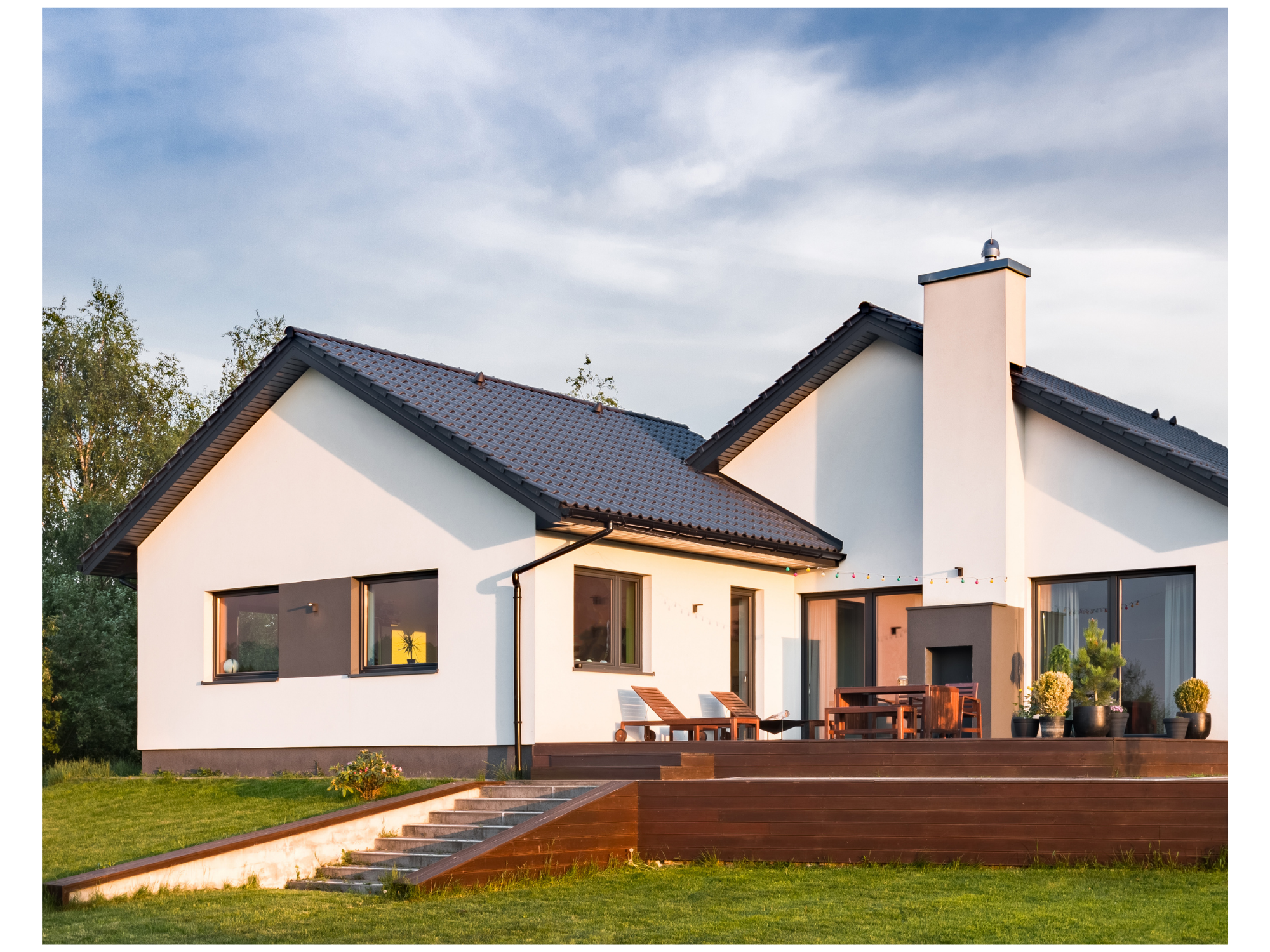 Modern two-story house with white walls, dark roof, and large windows, surrounded by a green lawn, trees, and outdoor furniture, under a partly cloudy sky.