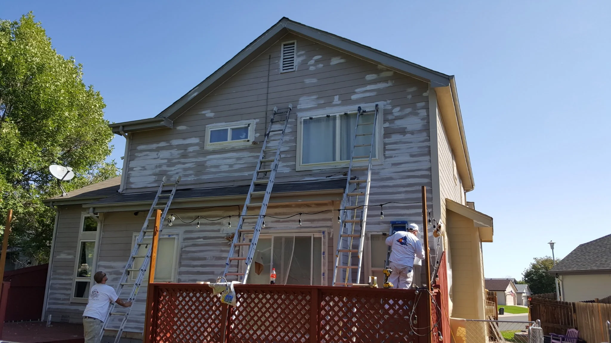 Workers applying siding to the second story of a house with ladders and tools, with sunlight and blue sky overhead.