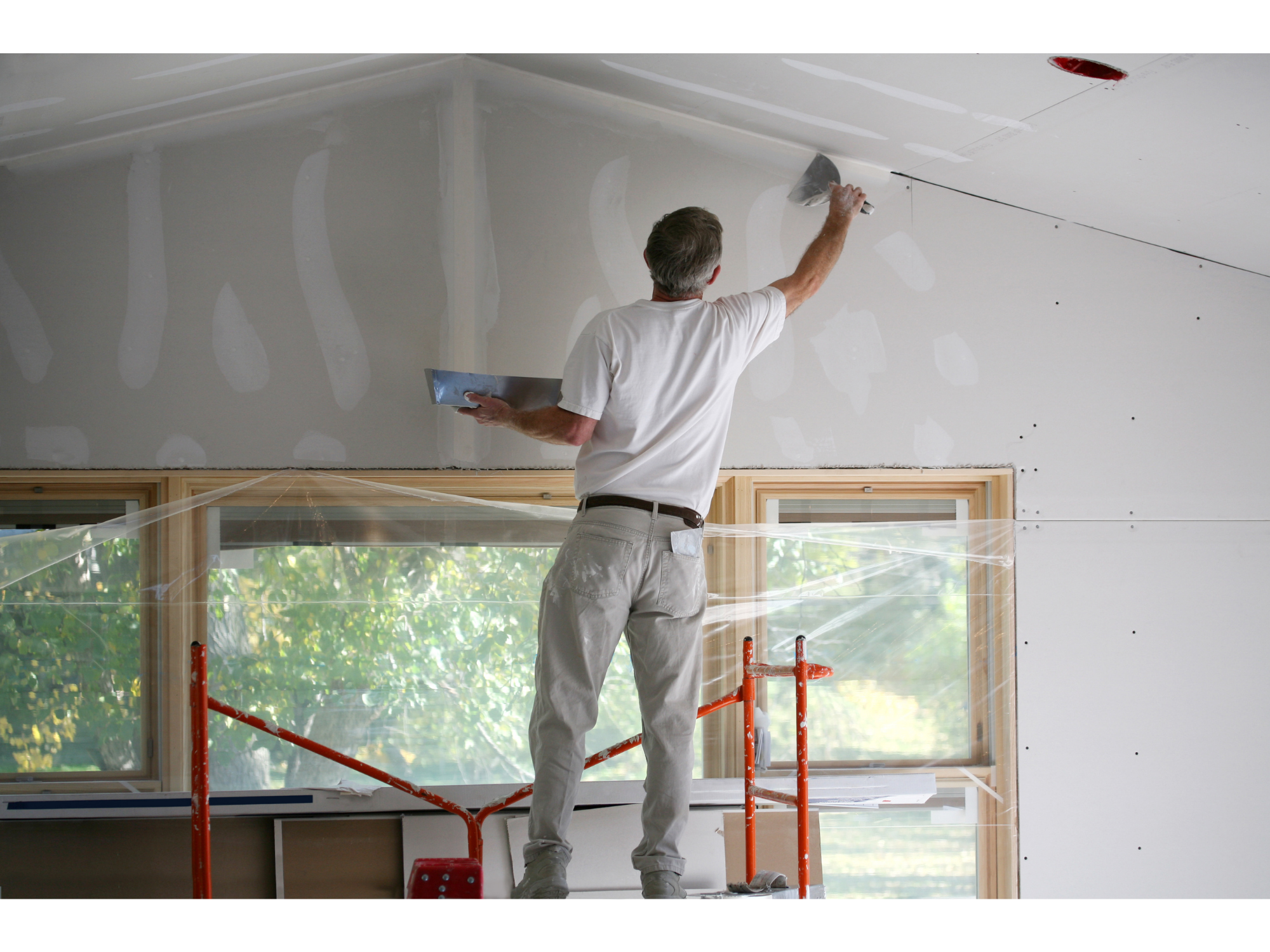 A person working on drywall installation or finishing in a room, standing on scaffolding, with a trowel in hand, applying joint compound to the ceiling, with a large window and natural light in the background.