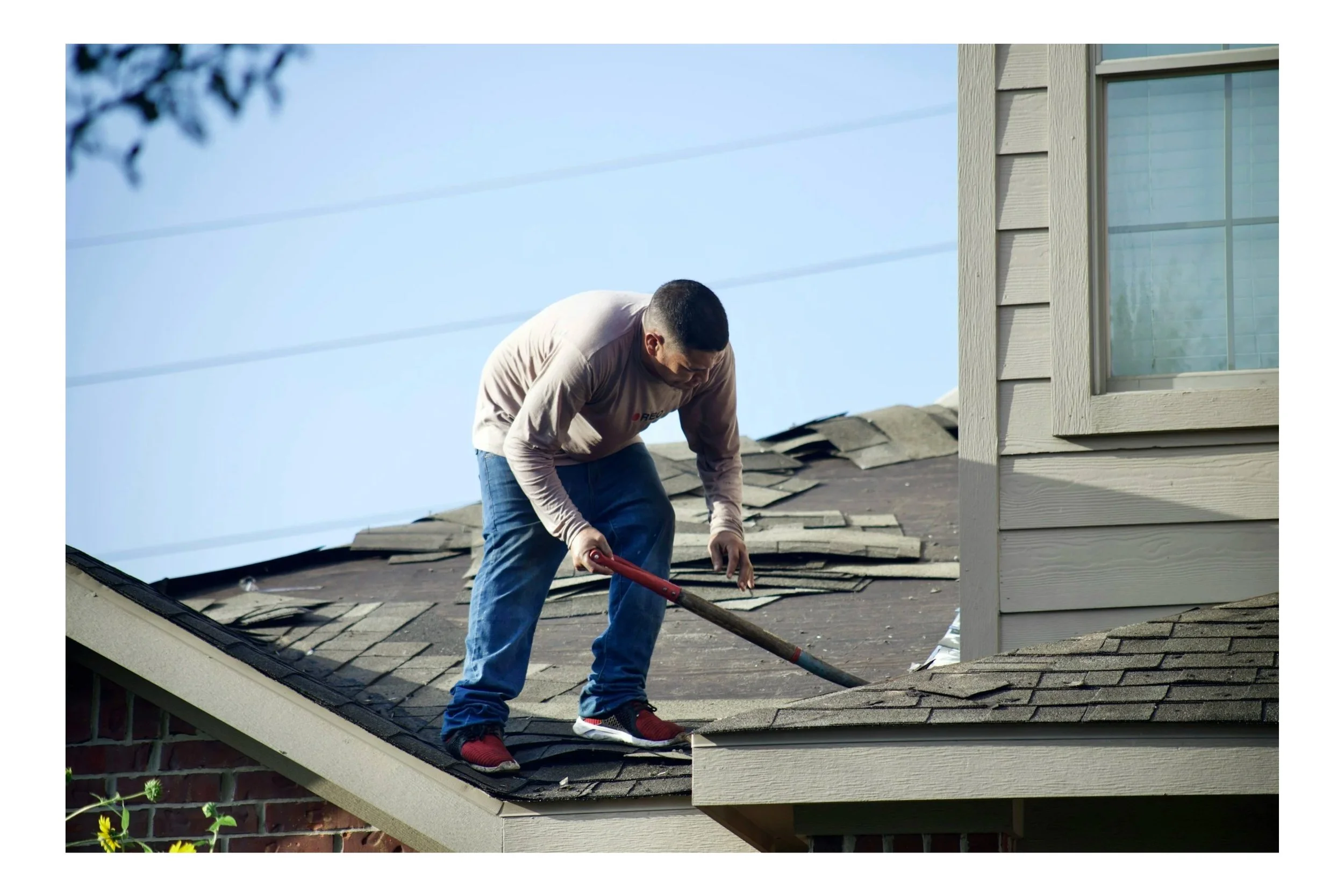 A man working on the roof of a house, using a tool on the shingles near the gutter, with a house window visible on the right and power lines in the background.