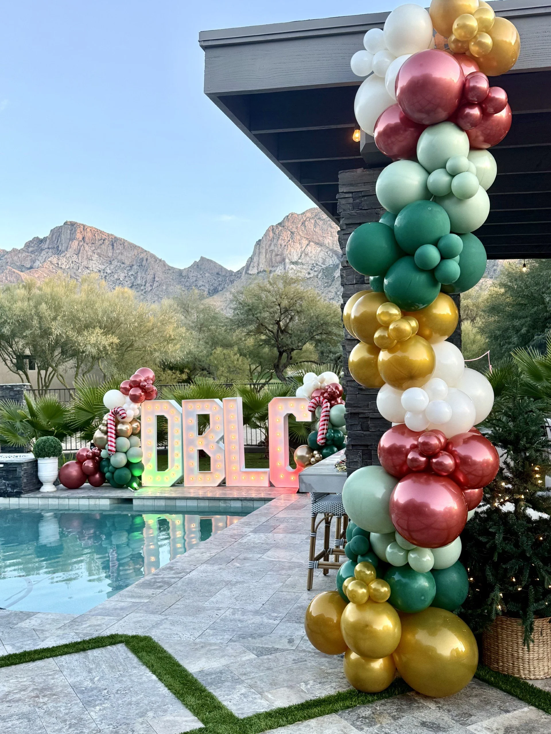 Outdoor poolside party decor with a large balloon arrangement in gold, white, red, green, and pink, and illuminated marquee letters spelling 'LOVE' in the background, with mountainous scenery in the distance.