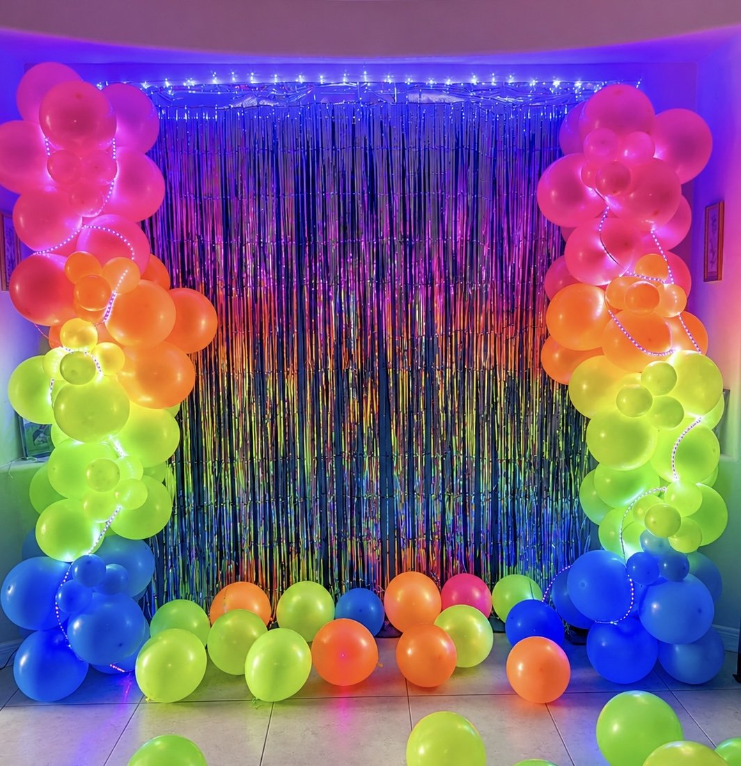 Colorful balloon arch with pink, orange, yellow, green, and blue balloons around a shimmering metallic backdrop at a festive party.