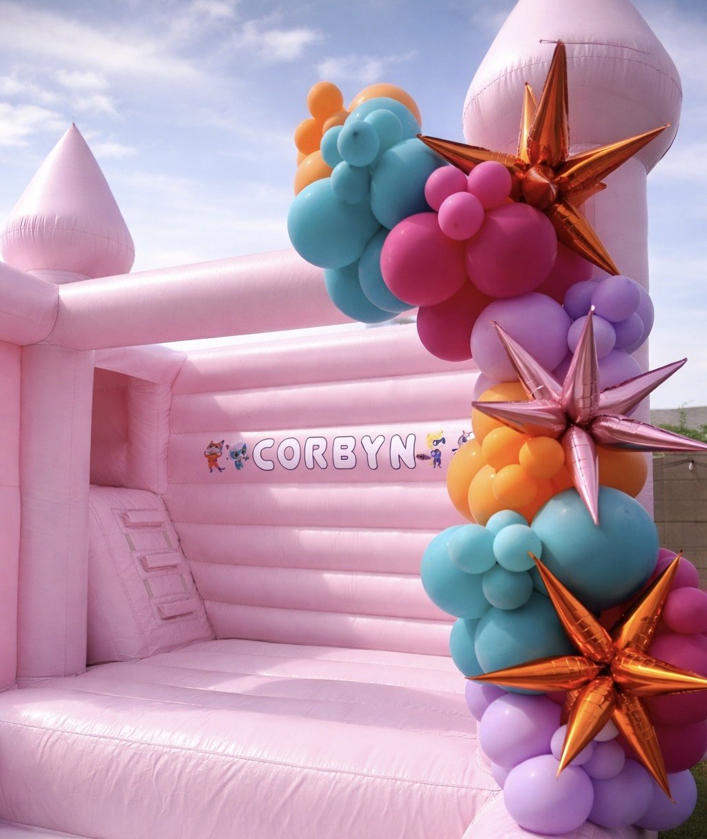 Pink inflatable bounce house decorated with colorful balloons and gold star-shaped balloons, with the name 'Corbyn' written on the side.