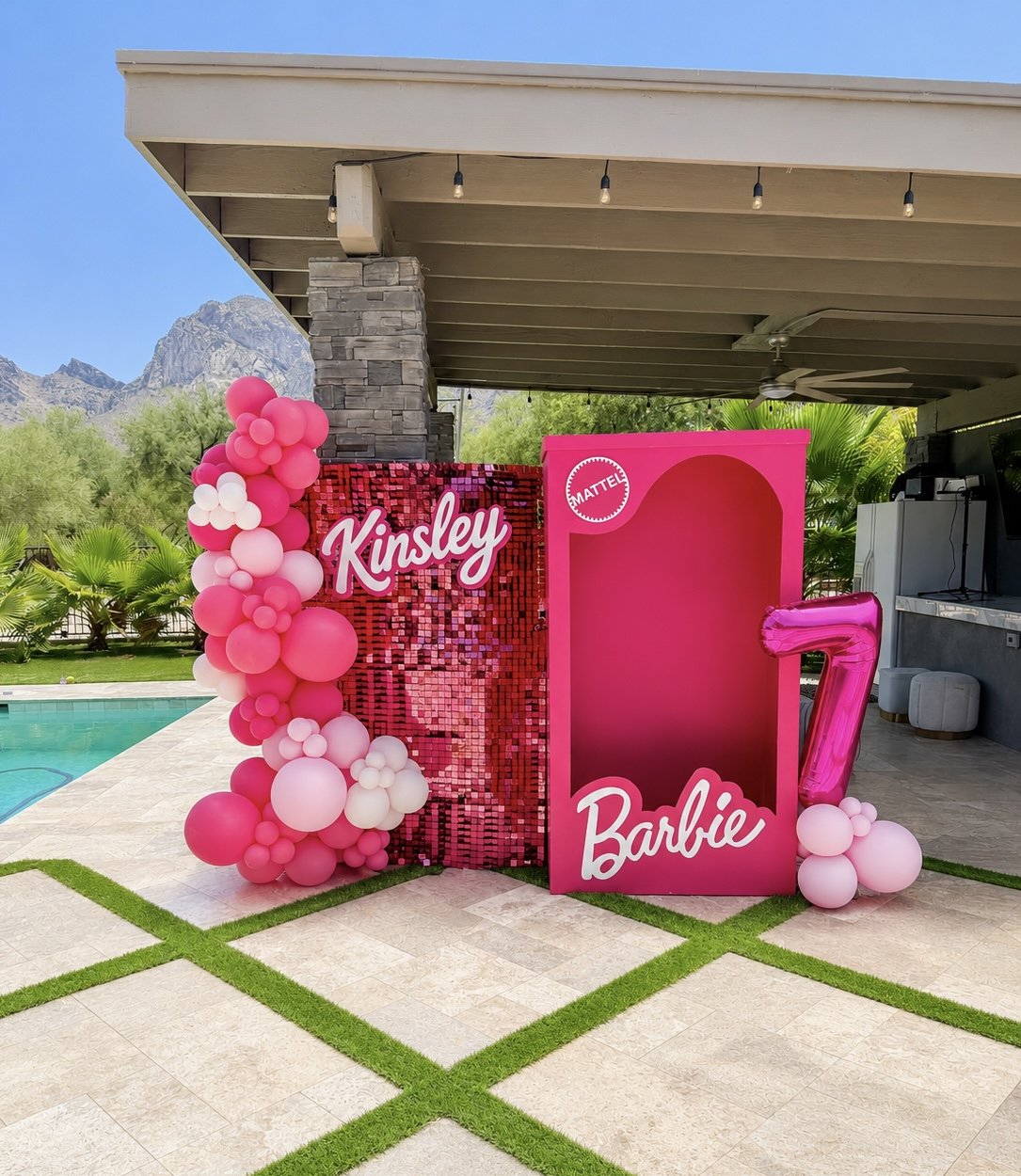 Decorative backdrop featuring pink and white balloons, with the names 'Kinsley' and 'Barbie' in pink script, a pink 'MATTE' circle, and a large pink number 7, set up outdoors near a swimming pool with mountains and greenery in the background.