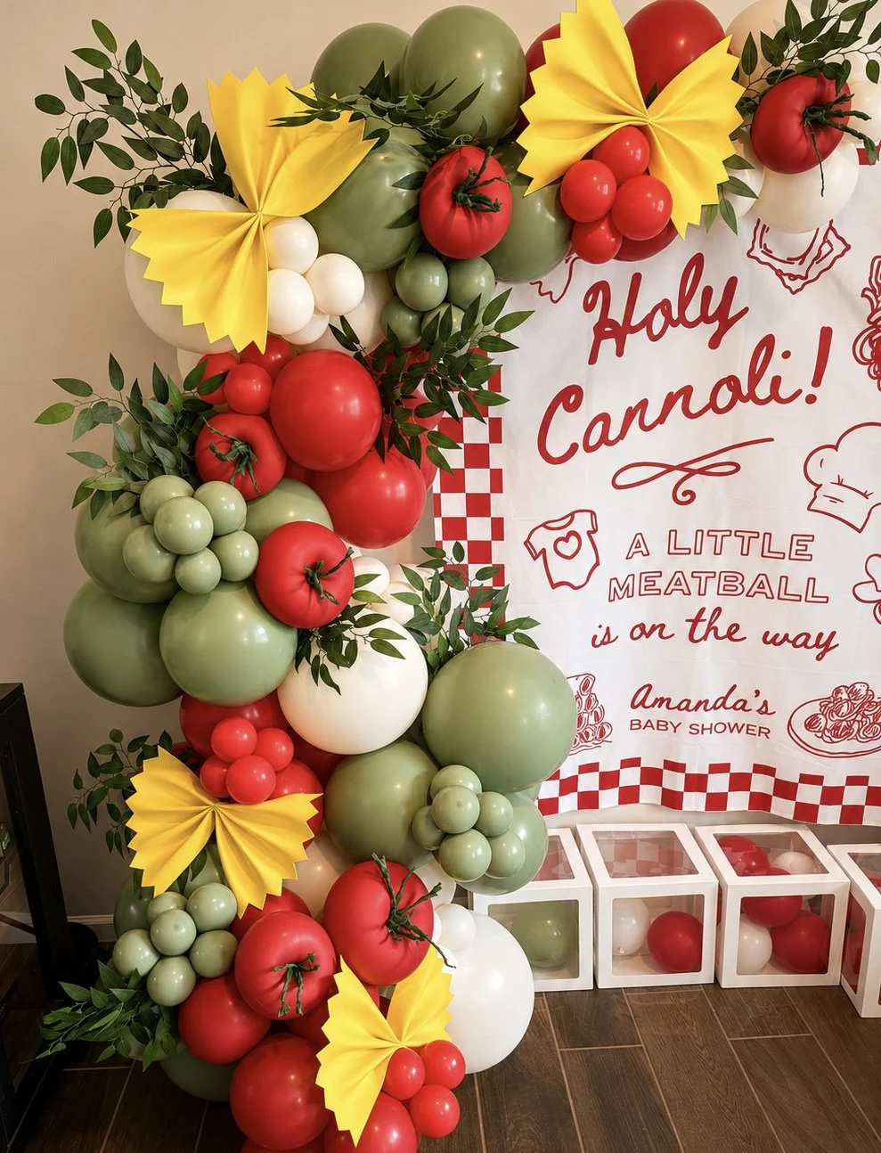 Decorative balloon arrangement with red, white, and green balloons resembling tomatoes, decorated with green leaves and yellow paper decorations, set against a backdrop with text for a baby shower celebration.