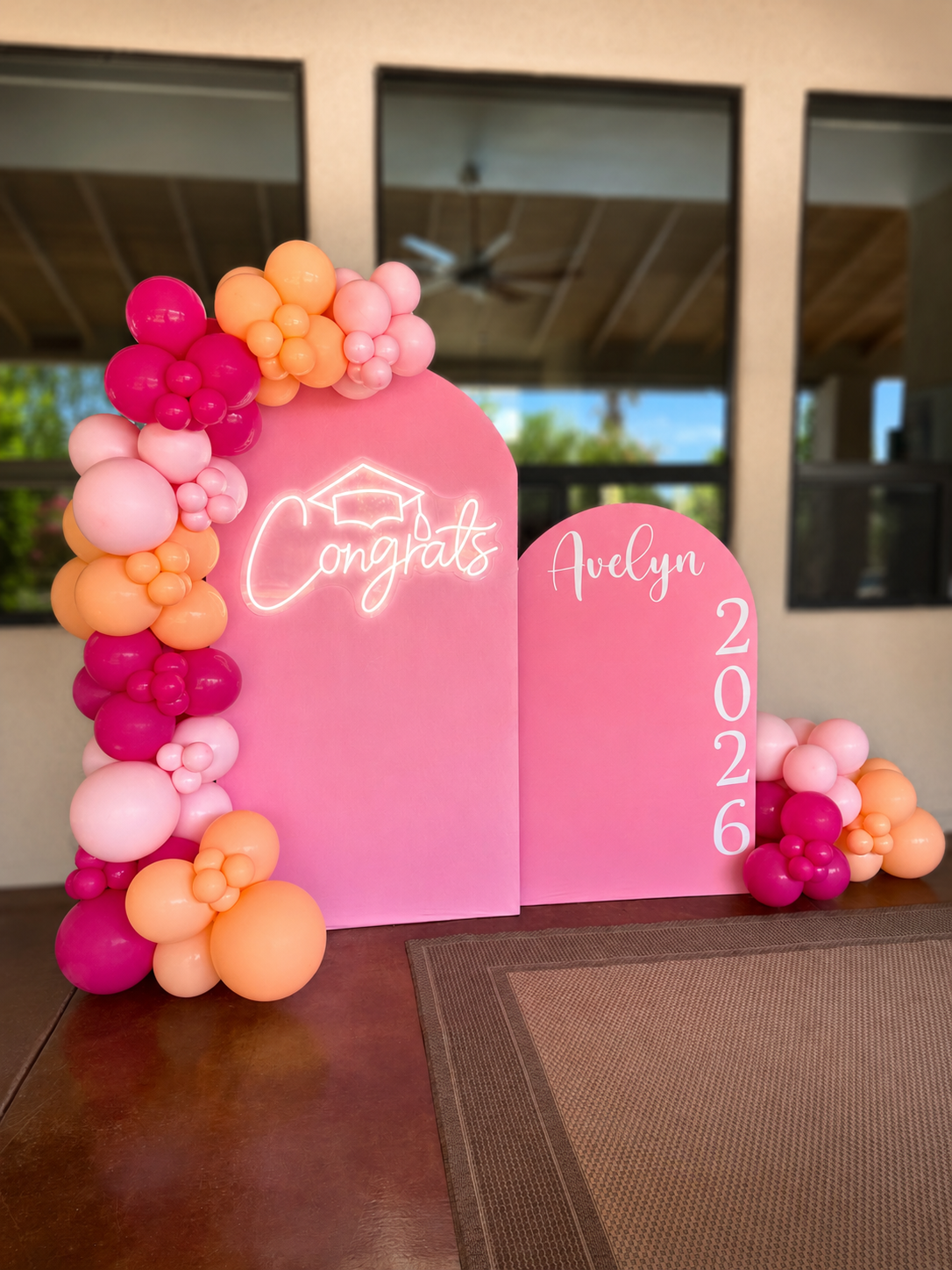 Pink display board decorated with balloons in shades of pink, peach, and coral with neon sign reading 'Congrats' and white text 'Avelyn 2026'.