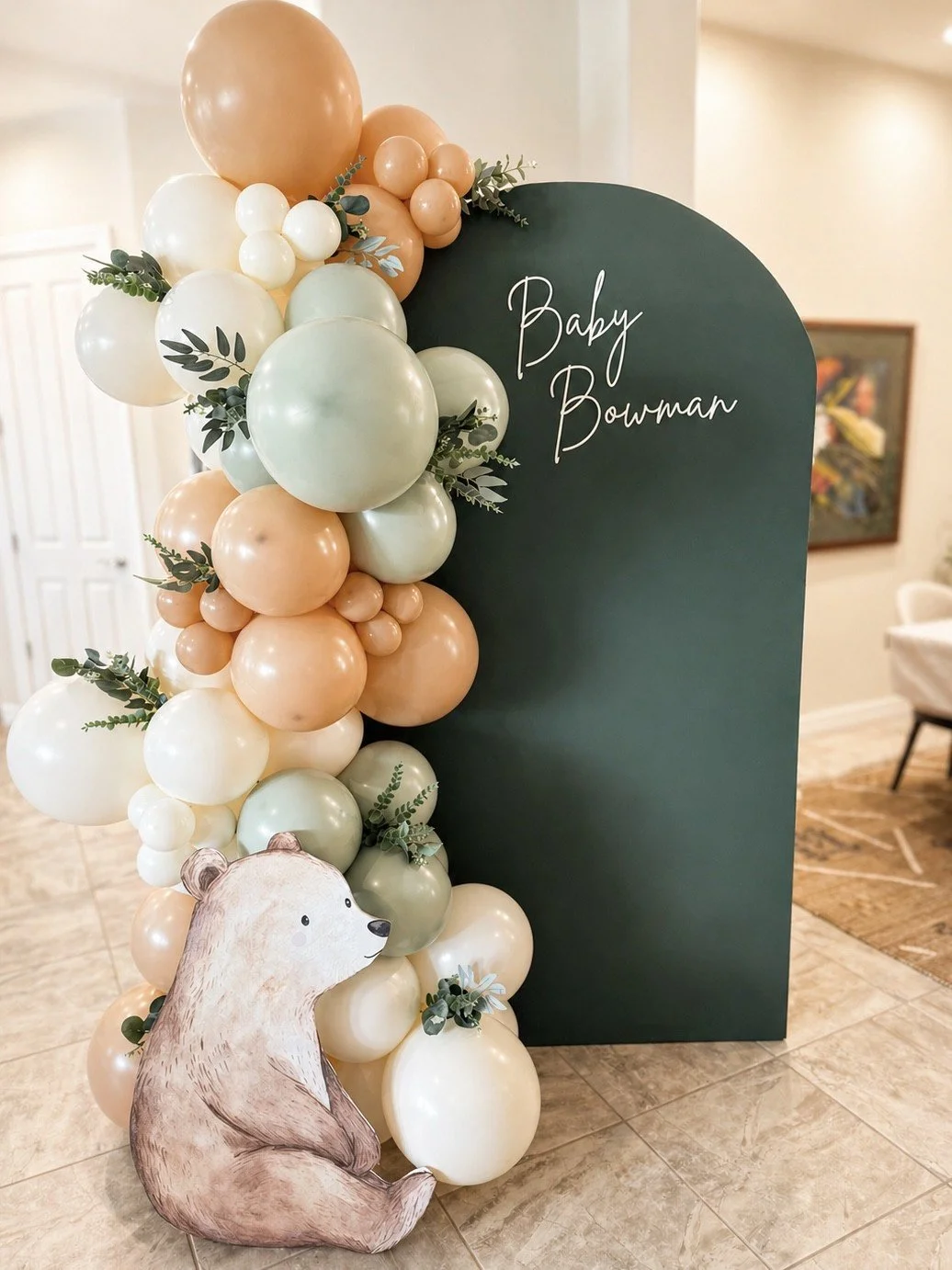 Decorative display with a dark green arch that has ‘Baby Bowan’ written on it, surrounded by a cascading arrangement of pastel-colored balloons and greenery, and a paper cutout of a sitting bear in front.