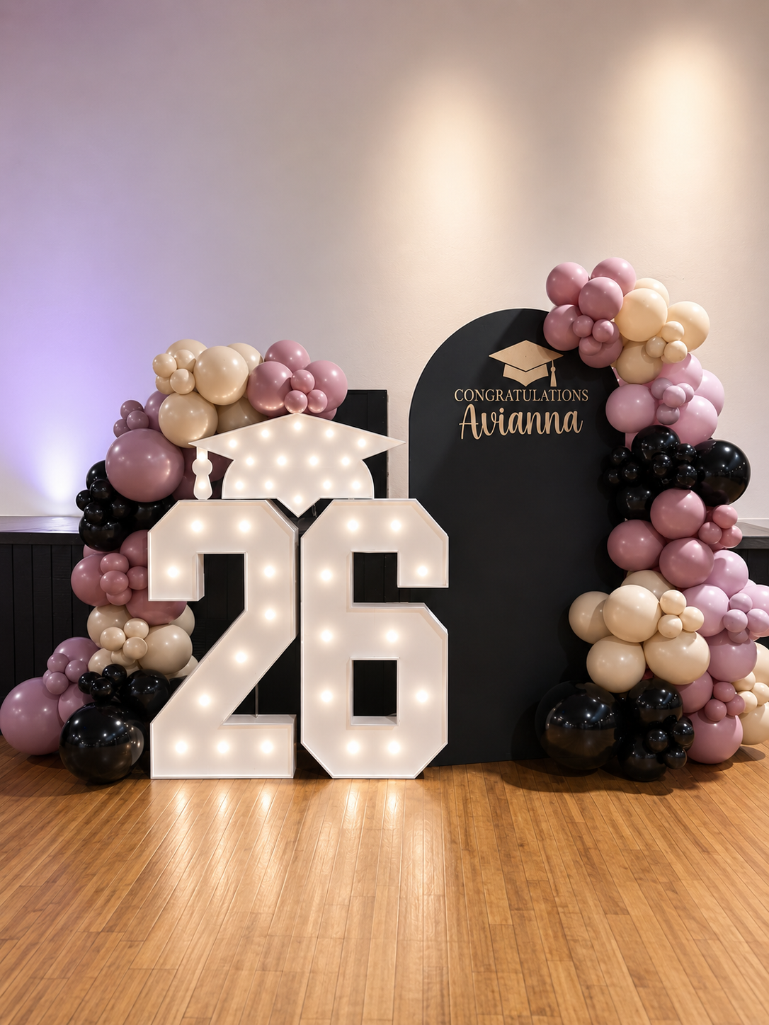 Decorations for a graduation celebration featuring a large illuminated number 26, pink, black, and beige balloons, and a black backdrop with a graduation cap graphic and the inscription 'Congratulations Avianna'.
