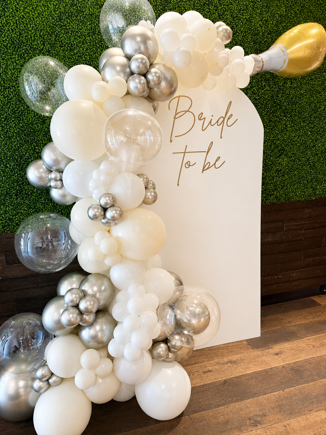 Decorative balloon arrangement with white, silver, and transparent balloons next to a sign that reads 'Bride to be', with a green hedge background and wooden floor.