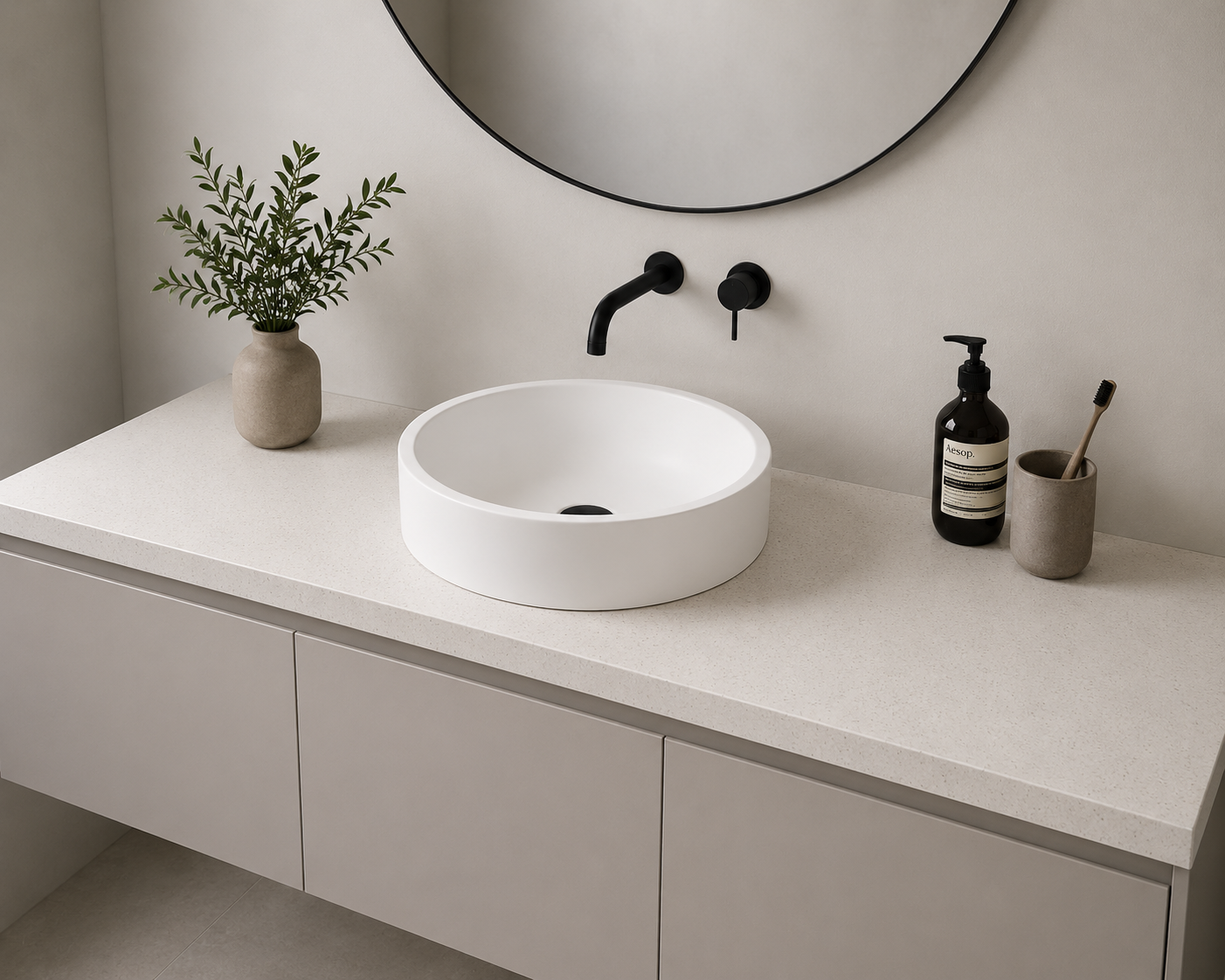 Minimalist bathroom vanity with round white vessel sink, black wall-mounted faucet, and a large mirror above. The counter has a beige vase with greenery, a black soap dispenser, and a beige cup with a toothbrush. Formed Solid Surface