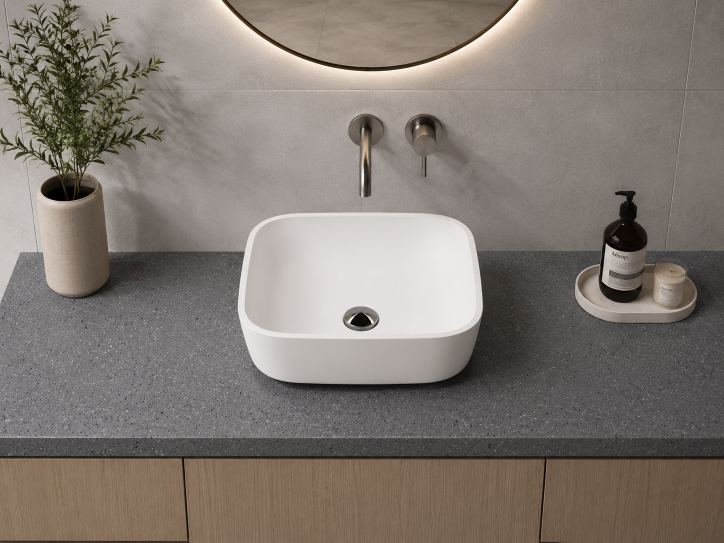 Modern bathroom countertop with a white vessel sink, a potted plant on the left, soap dispenser and candle on the right, and a round mirror above. Formed Solid Surface