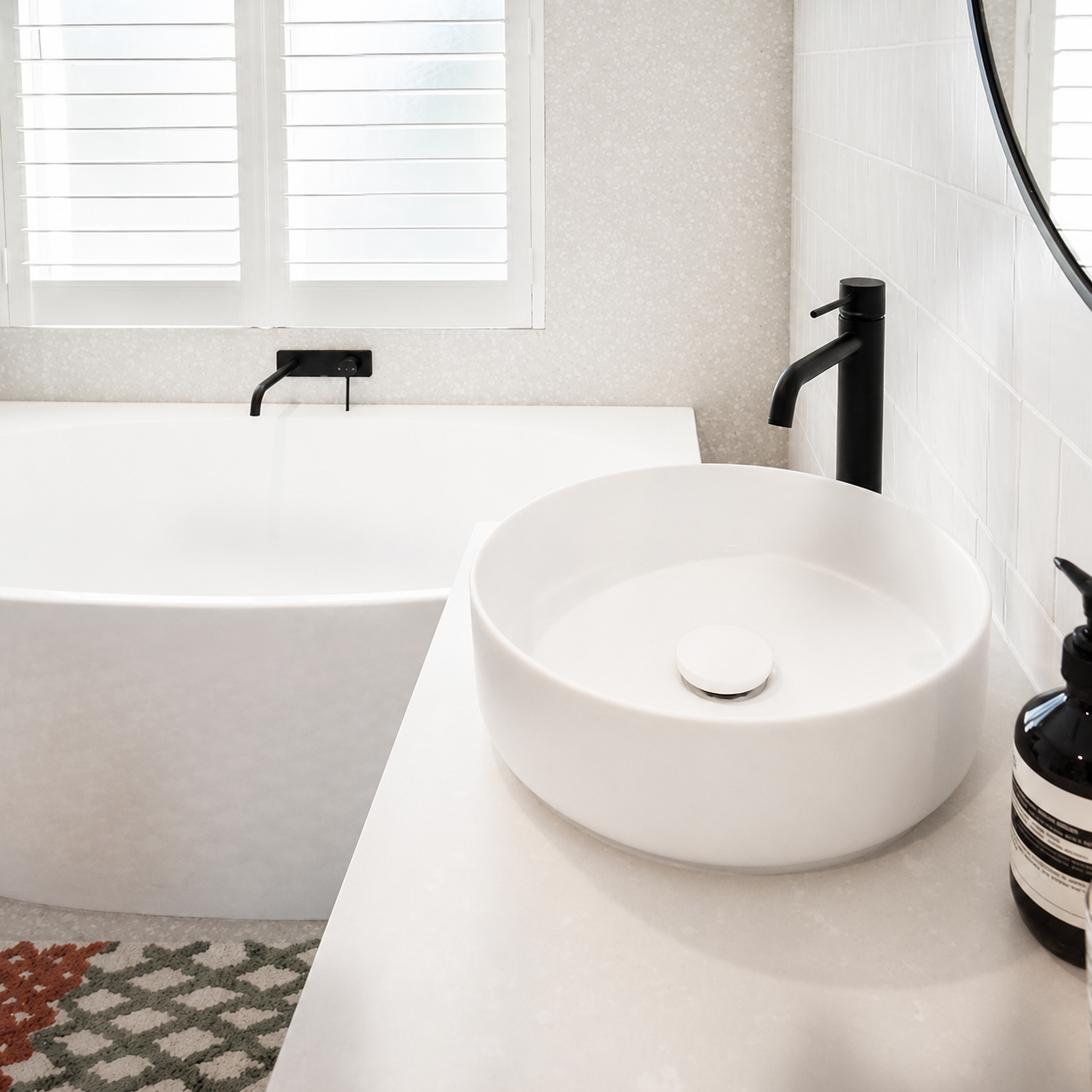 Modern bathroom with a white countertop, round white sink, black faucet, black soap dispenser, bathtub, window with shutters, and a patterned rug. Formed Solid Surface