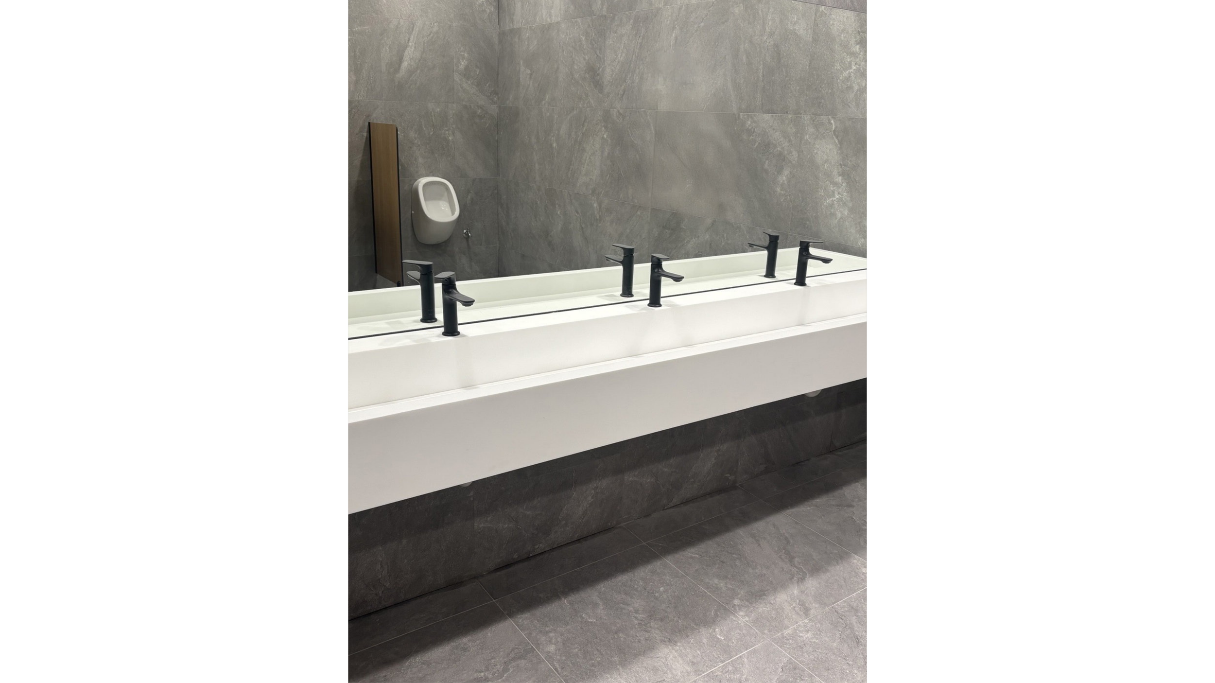 Public restroom with gray tiled walls and floor, a row of four black faucets over a white trough sink, and a wall-mounted urinal with a wooden panel behind it. Formed Solid Surface