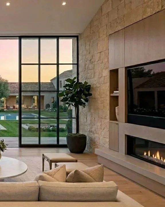 Living room with large window, potted plant, beige sofa, round coffee table, and a fireplace built into a wooden panel, with a view of a backyard with a swimming pool and houses.