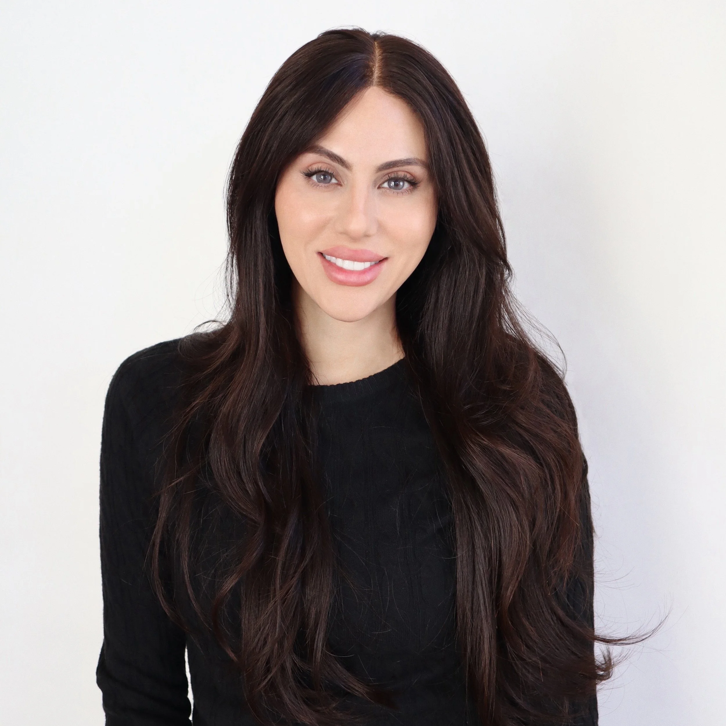Portrait of a woman with long dark hair, light makeup, wearing a black top, standing against a plain white background.