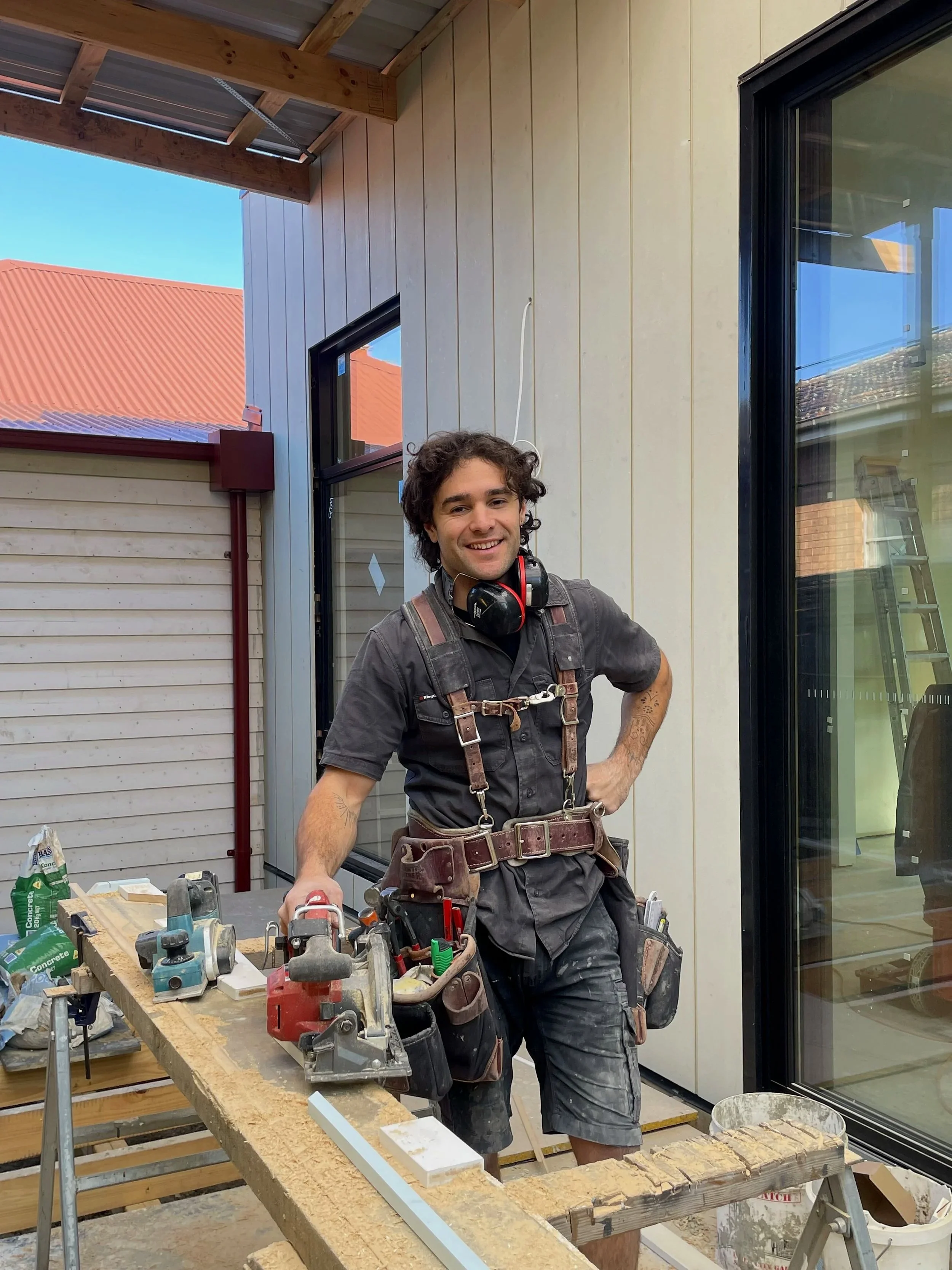 A man with curly hair and a smile, wearing a tool harness, stands in a construction workspace surrounded by tools and materials, indoors near a glass door and a window with a view of neighboring buildings with red roofs.