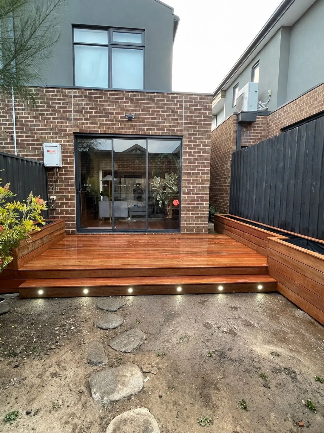 A partially constructed or renovated outdoor patio area with tools and equipment scattered on the wooden deck. The area is enclosed by glass doors and windows, with trees and a wooden fence visible outside. Light filters through the trees casting shadows on the deck.