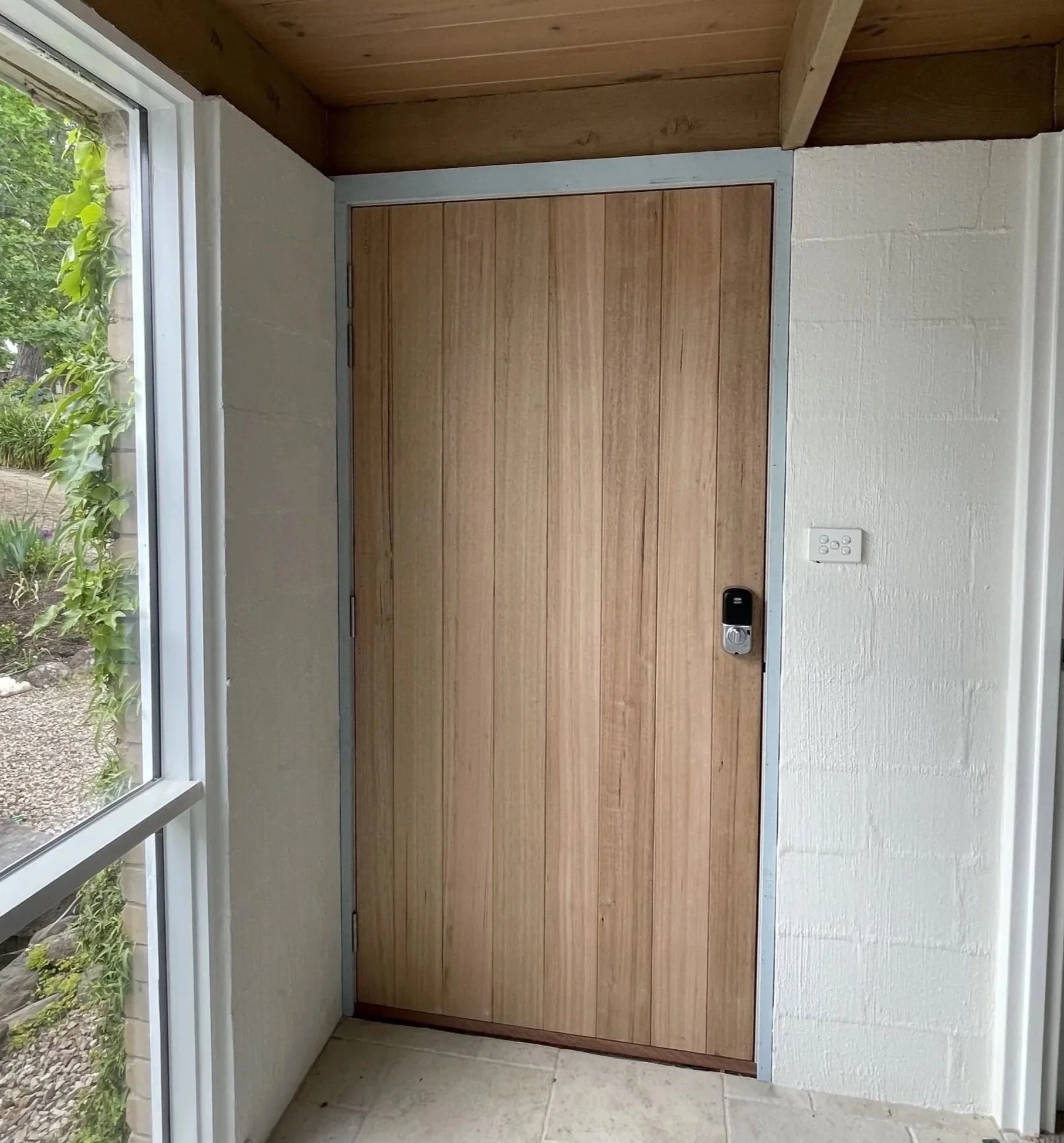 A wooden door installed in a brick wall with a doorbell and door handle. There is a green leafy plant on the right side of the door.