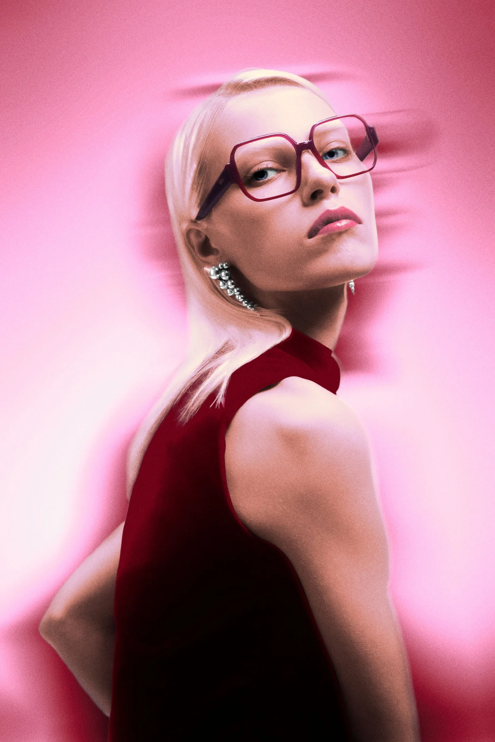 A woman with platinum blonde hair wearing large pink glasses and pearl earrings, dressed in a red sleeveless dress, against a pink background.