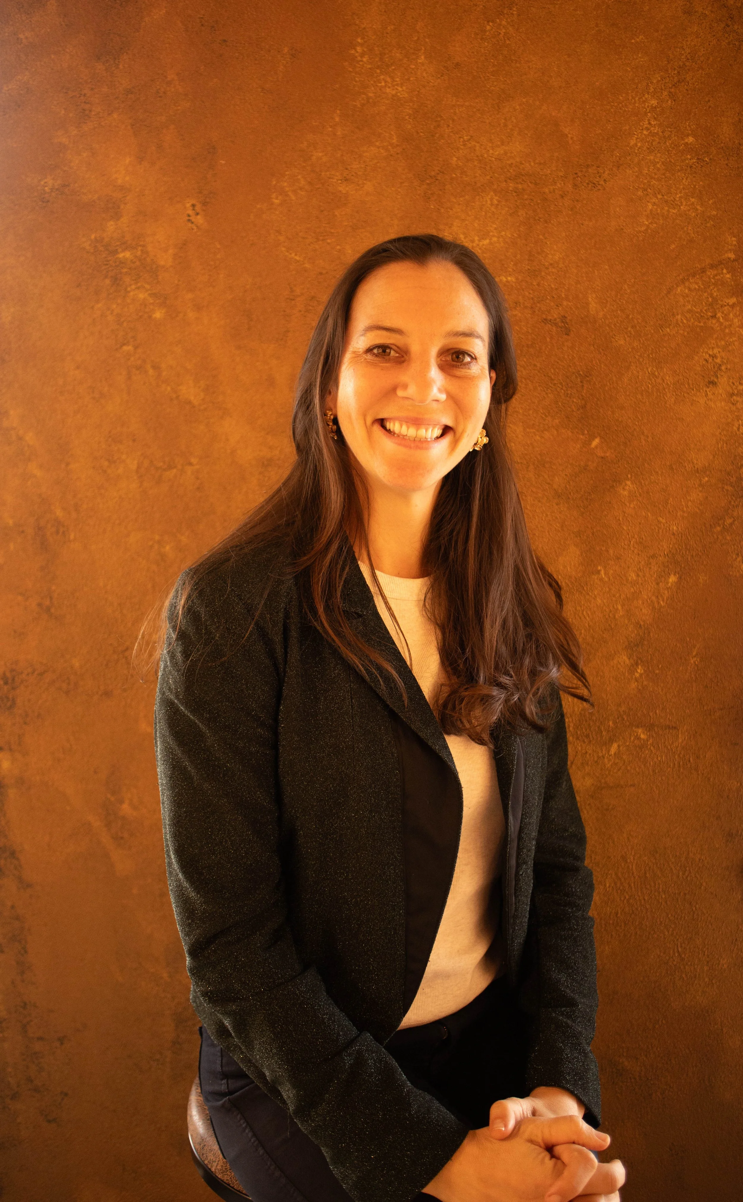A woman with long dark hair, wearing a black blazer and beige top, smiling at the camera against a textured brown background.