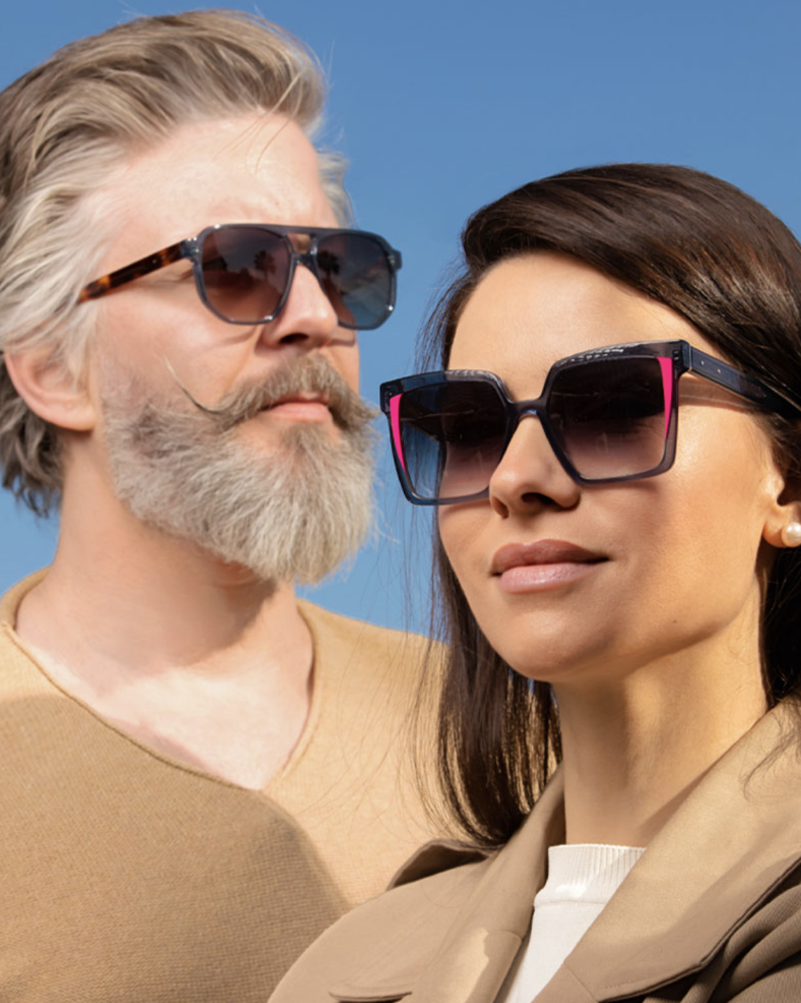 Close-up of a mature man with gray hair and beard wearing sunglasses, and a young woman with dark hair and earrings wearing large sunglasses, both against a blue sky.