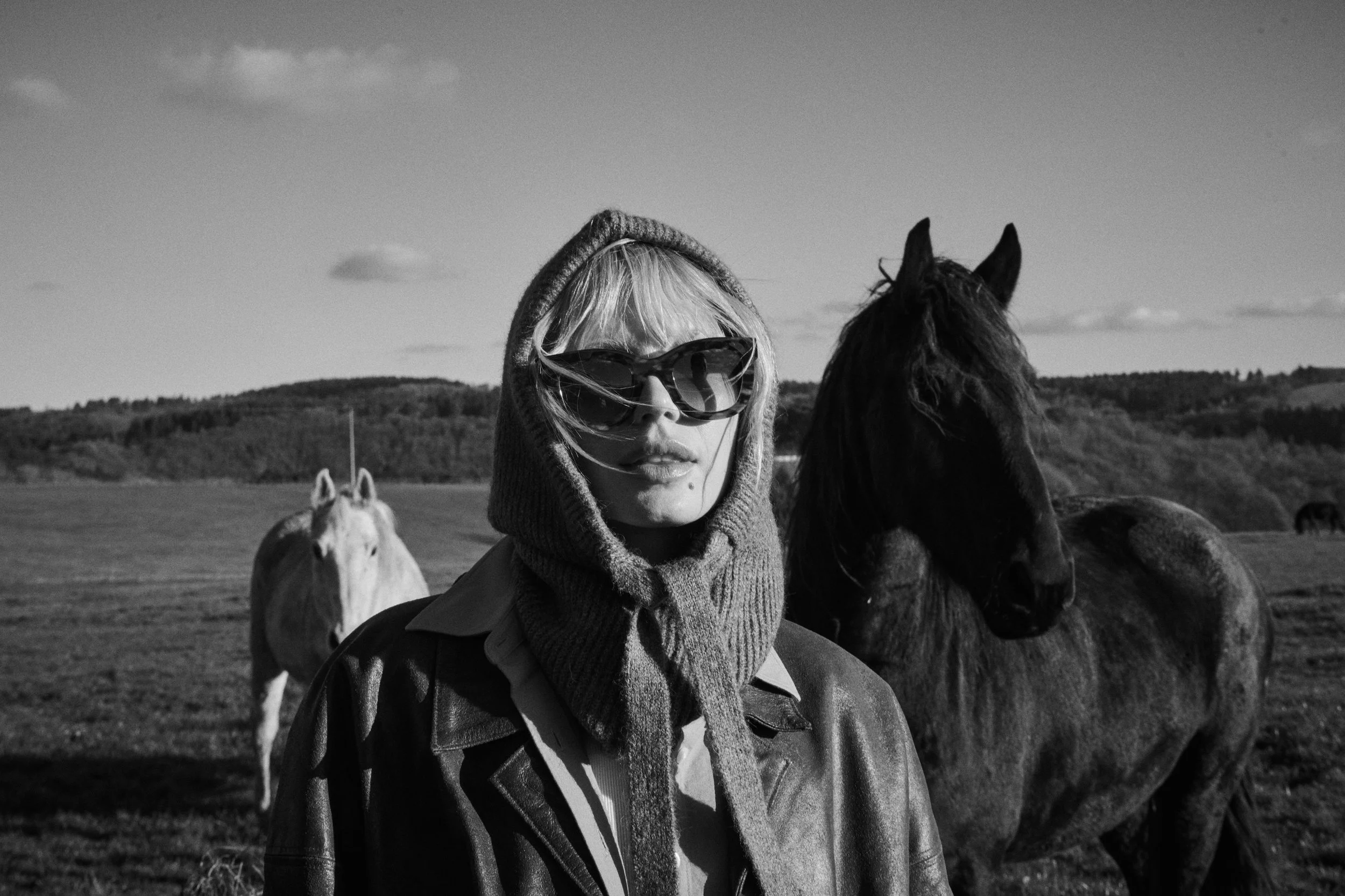A black and white photo of a woman in sunglasses with a hooded sweater, standing outdoors with horses in the background.